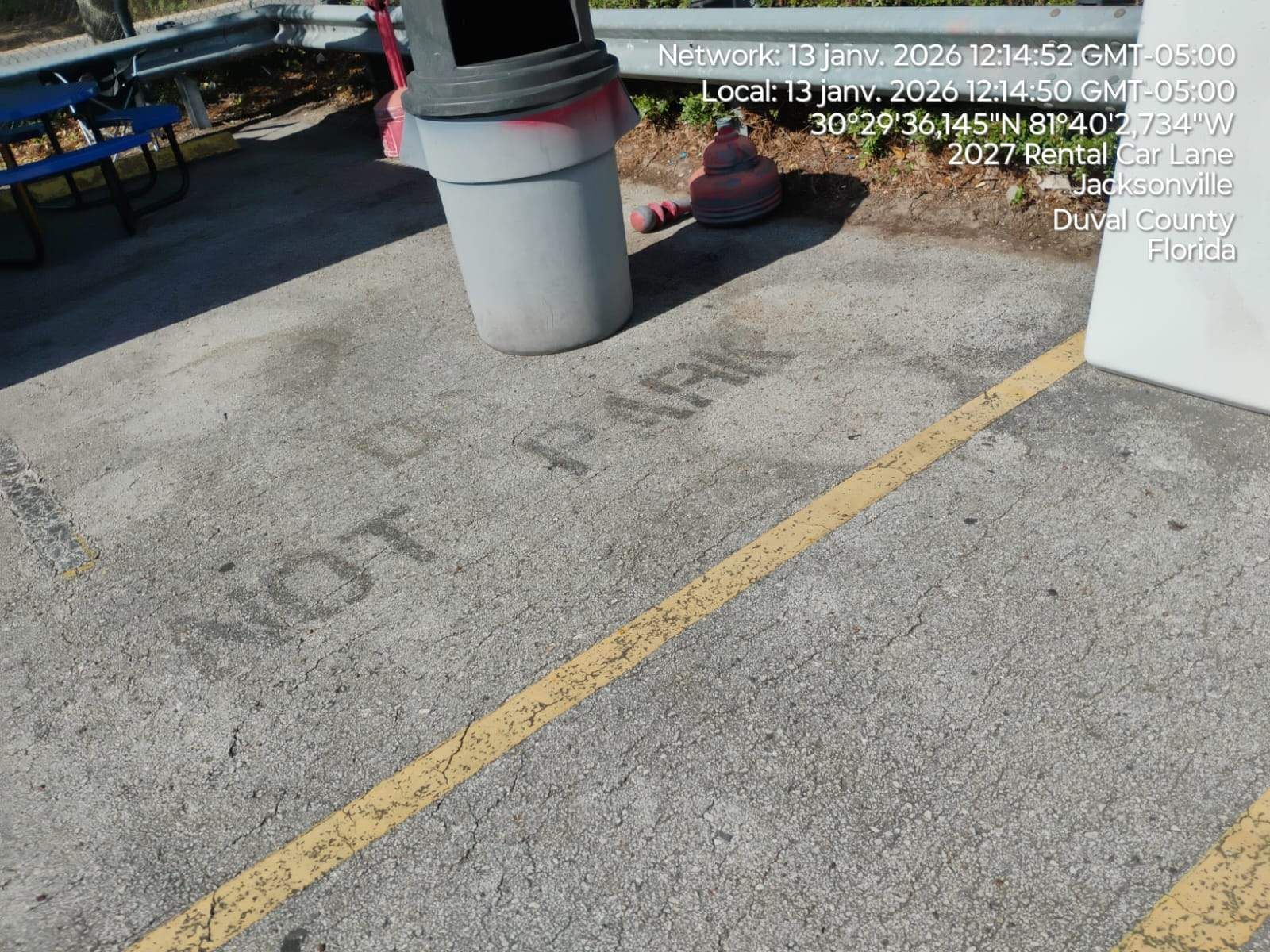Outdoor concrete parking area with yellow line, pole base, and a person sitting near a planter.