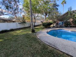 Backyard with pool, lawn, white fence, trees, and cloudy blue sky.