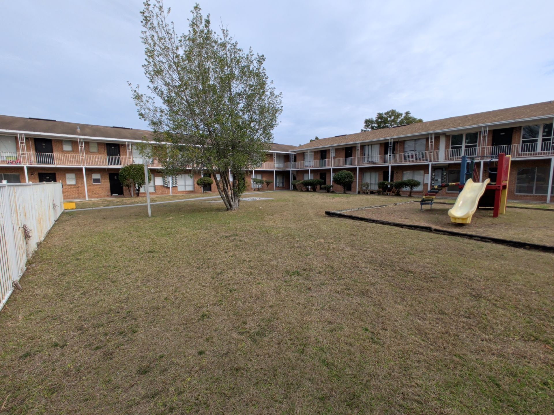 Courtyard with two-story brick apartment buildings, small playground, and a central tree under a cloudy sky.