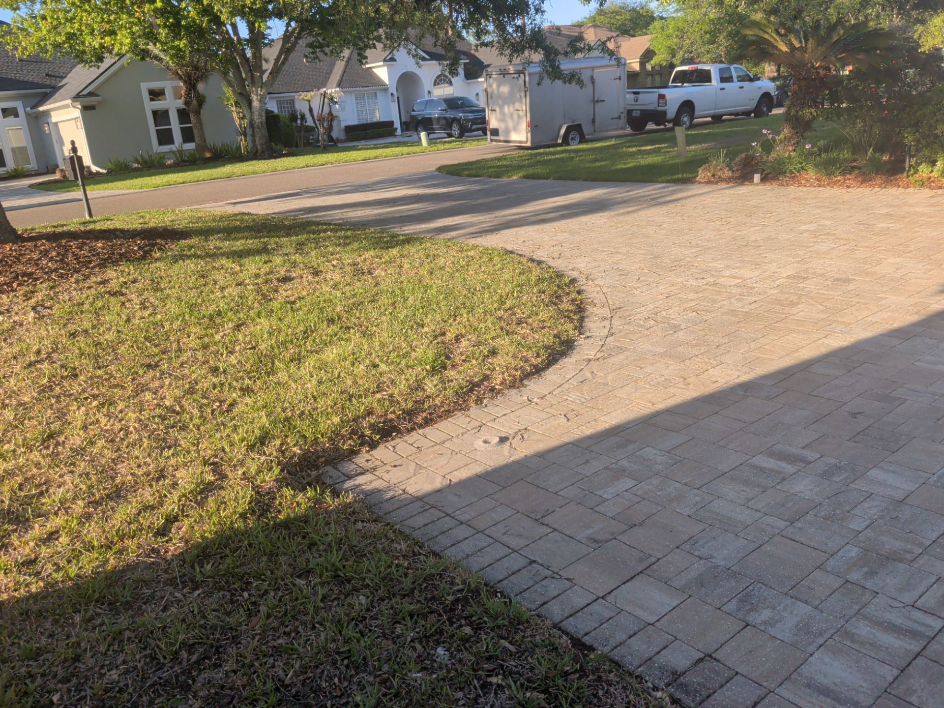 Residential driveway with brick pavers, curved grass edge, and houses with parked vehicles in the background