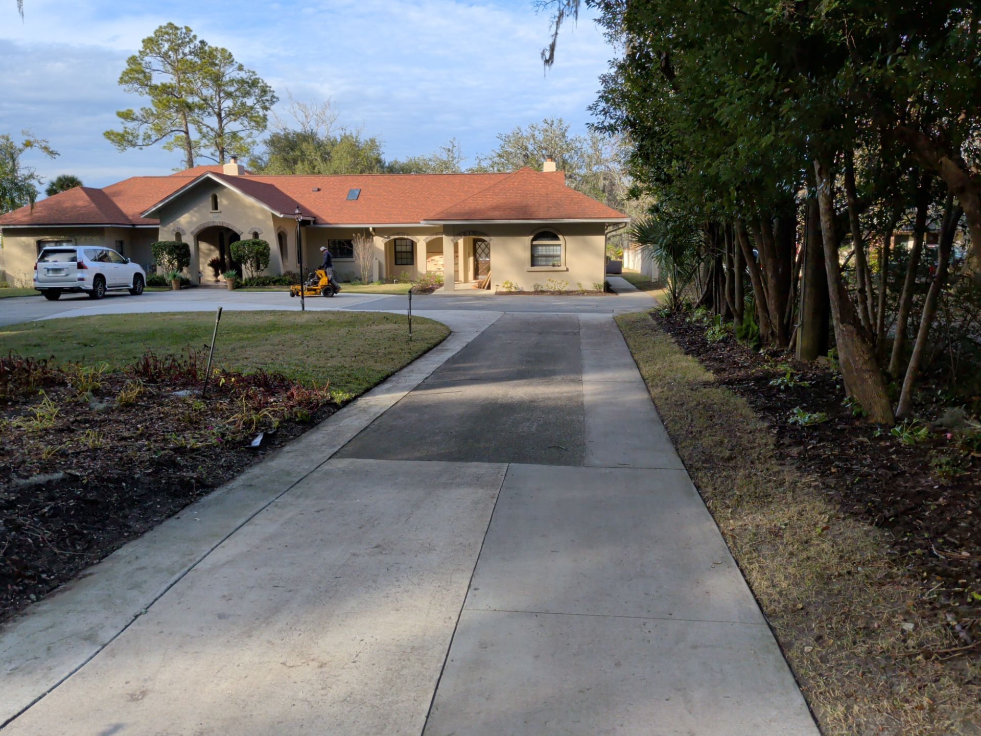 Long driveway leading to a tan house with a red tile roof and parked cars in front