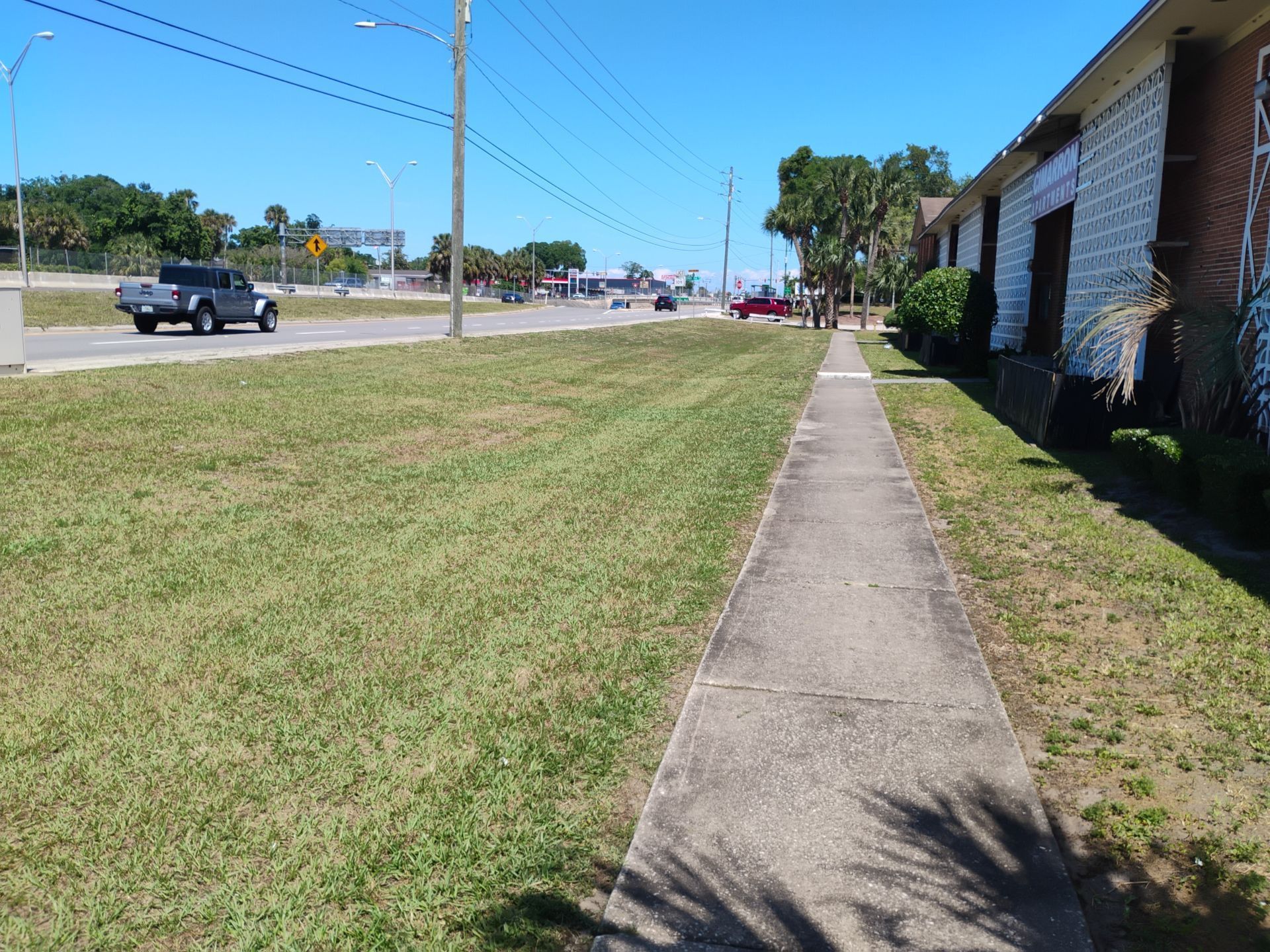 Sidewalk beside a grassy roadside under a clear blue sky, with cars on the street and a brick building nearby.