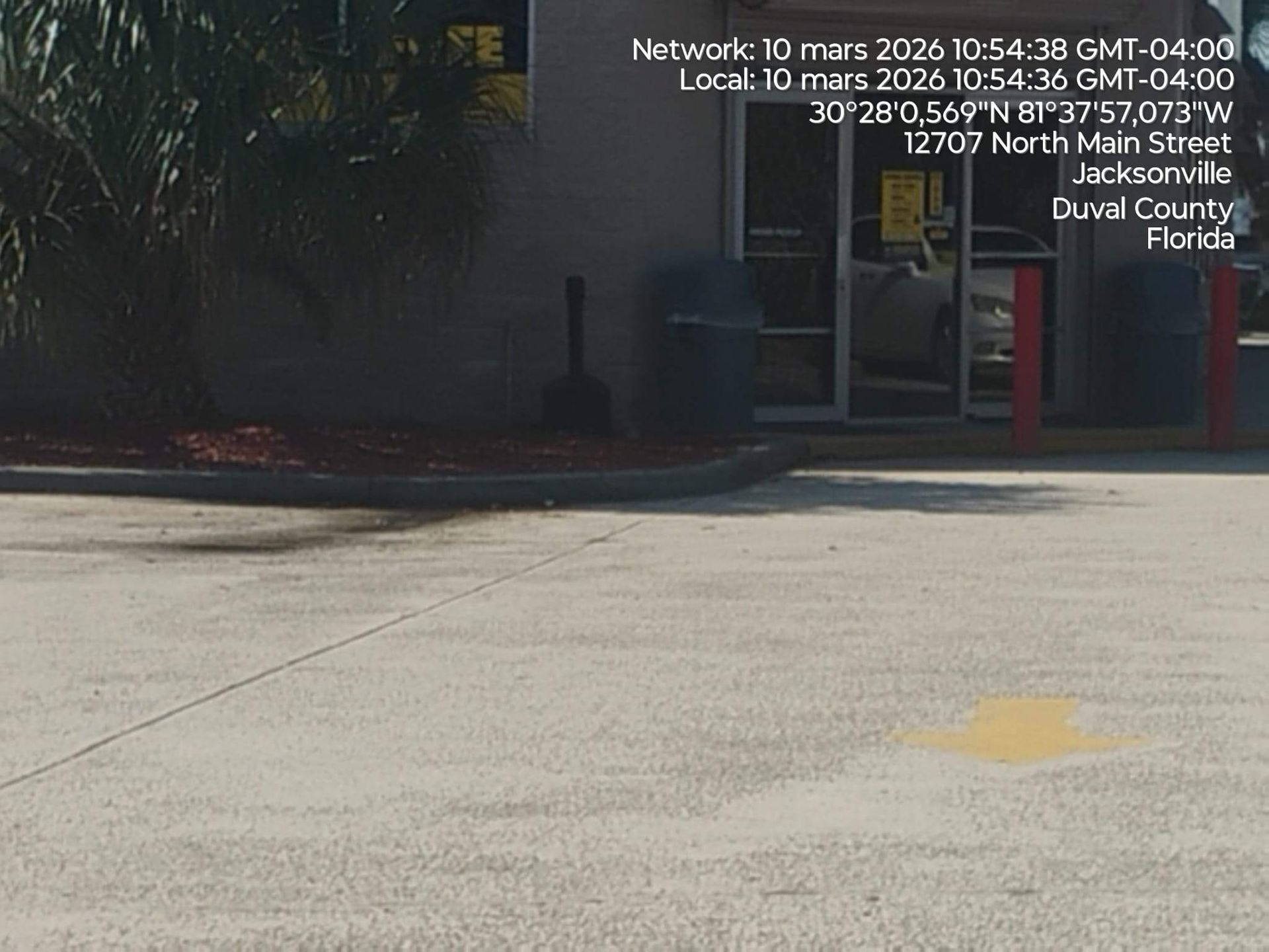 Sunlit parking lot outside a church entrance with glass doors and a red bollard, trees in the background