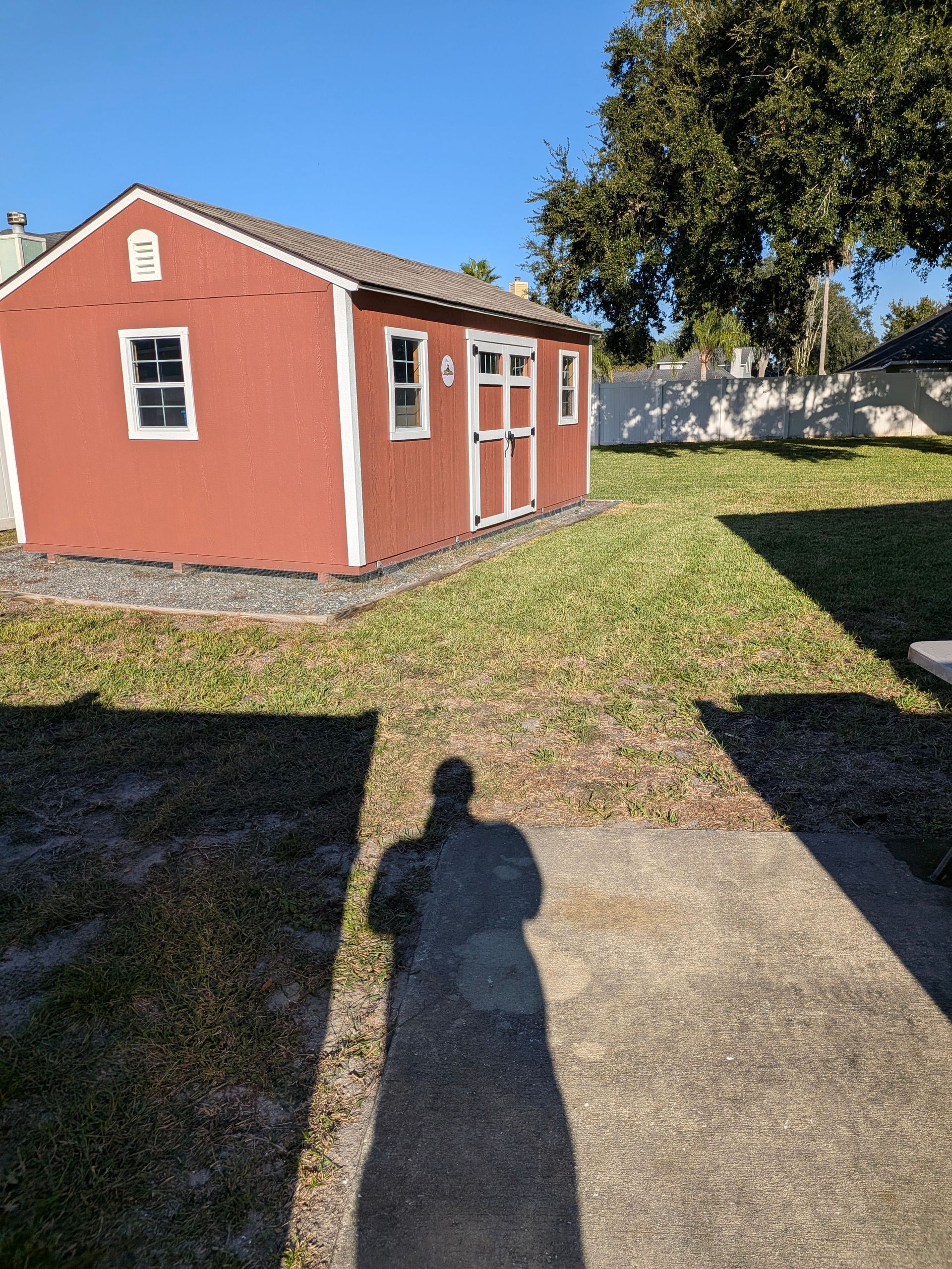 Red shed-style building in a grassy yard with a concrete path and long shadow in the foreground.