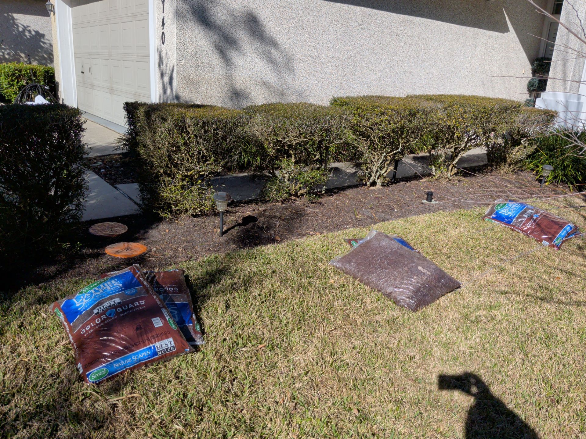 Bags of mulch on a lawn near a trimmed hedge and a house. Shadow of a person is visible.