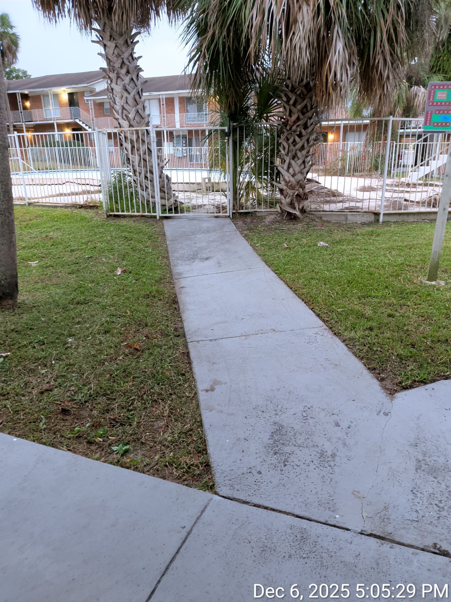 Concrete walkway through grassy yard toward a fenced apartment courtyard with palm trees and a pool in the background