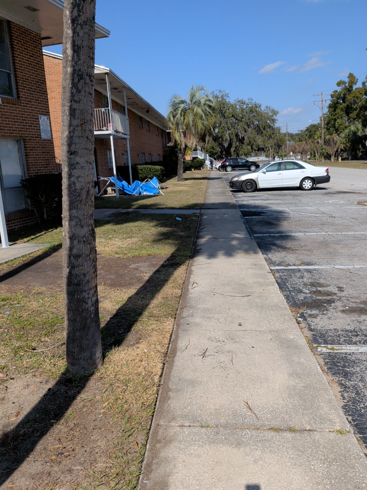 Sidewalk next to a brick building and parking lot with a car on a sunny day.