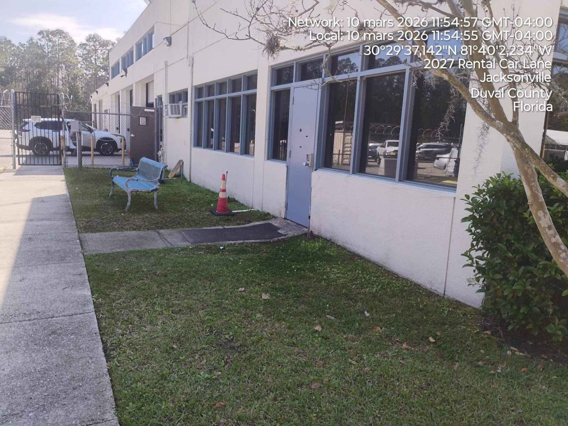 Apartment building exterior with sidewalk, grass, windows, and a blue bench near the entrance