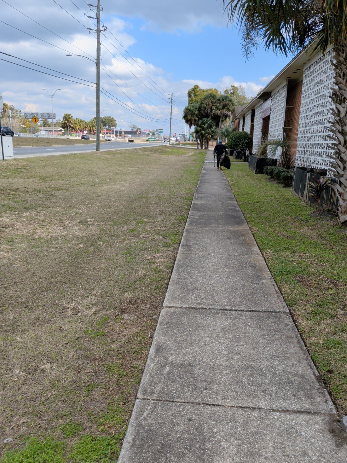 Sidewalk next to grass and a road, person walking, low-rise brick building on right, sunny day.