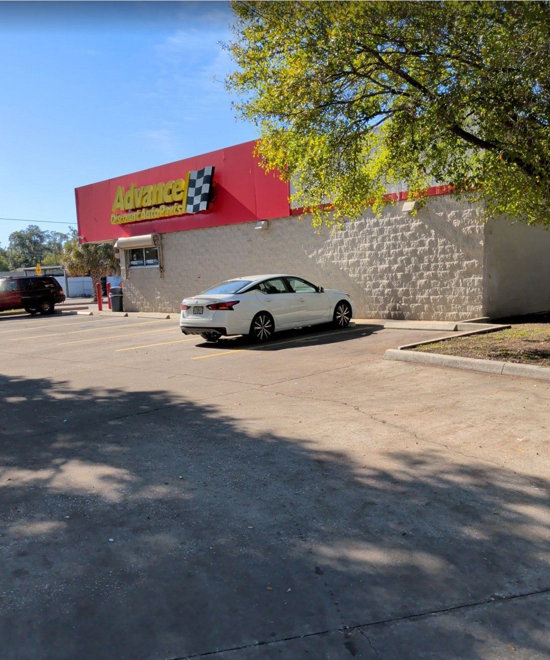 Parking lot outside a red storefront, with a silver car parked near a stone wall under a leafy tree.