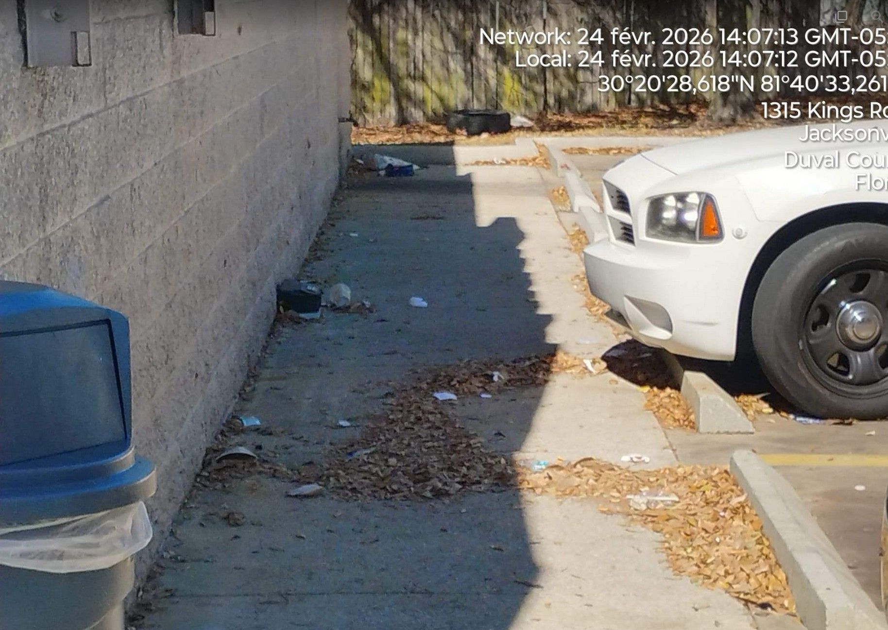 Narrow alley beside a white pickup truck, with scattered leaves and a blue trash bin along a concrete wall.
