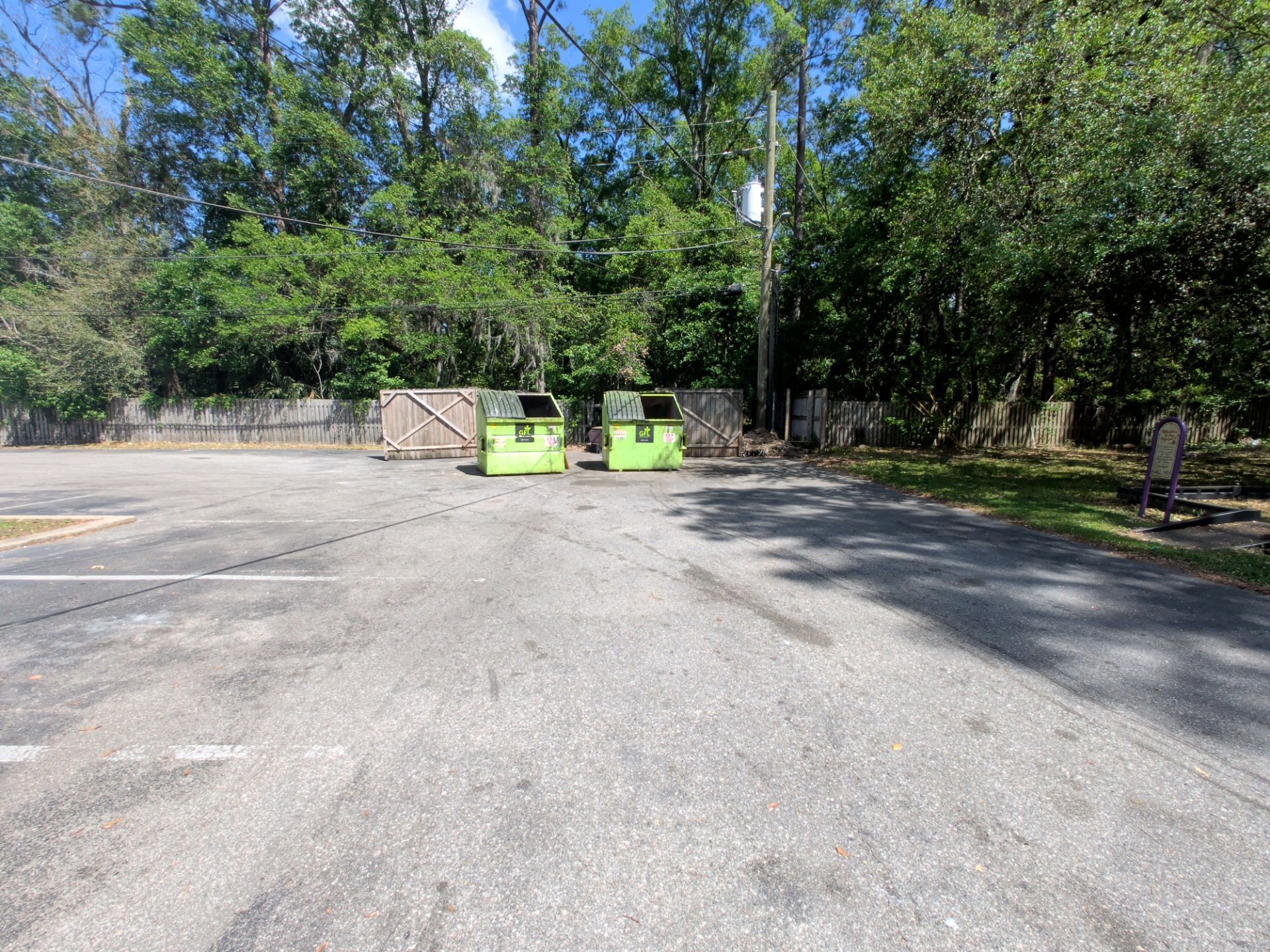 Two green dumpsters beside a wooden gate in a sunny paved parking area, with trees in the background