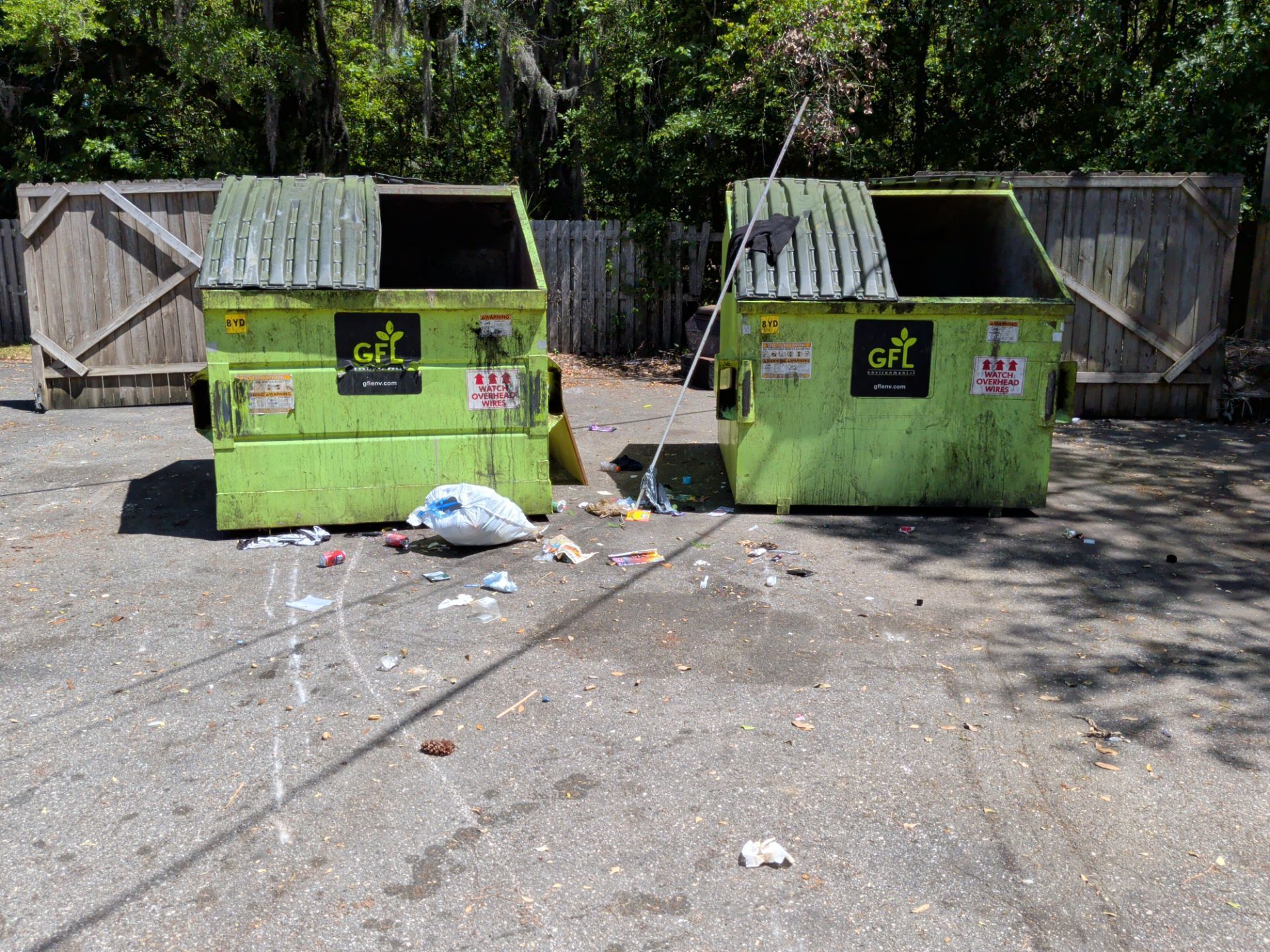 Two green dumpsters on a litter-strewn asphalt lot near a fence and trees.