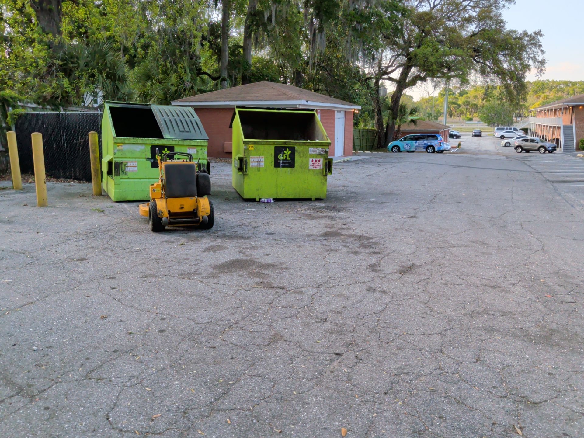 Green dumpsters and a yellow forklift in a gravel lot near trees and small buildings