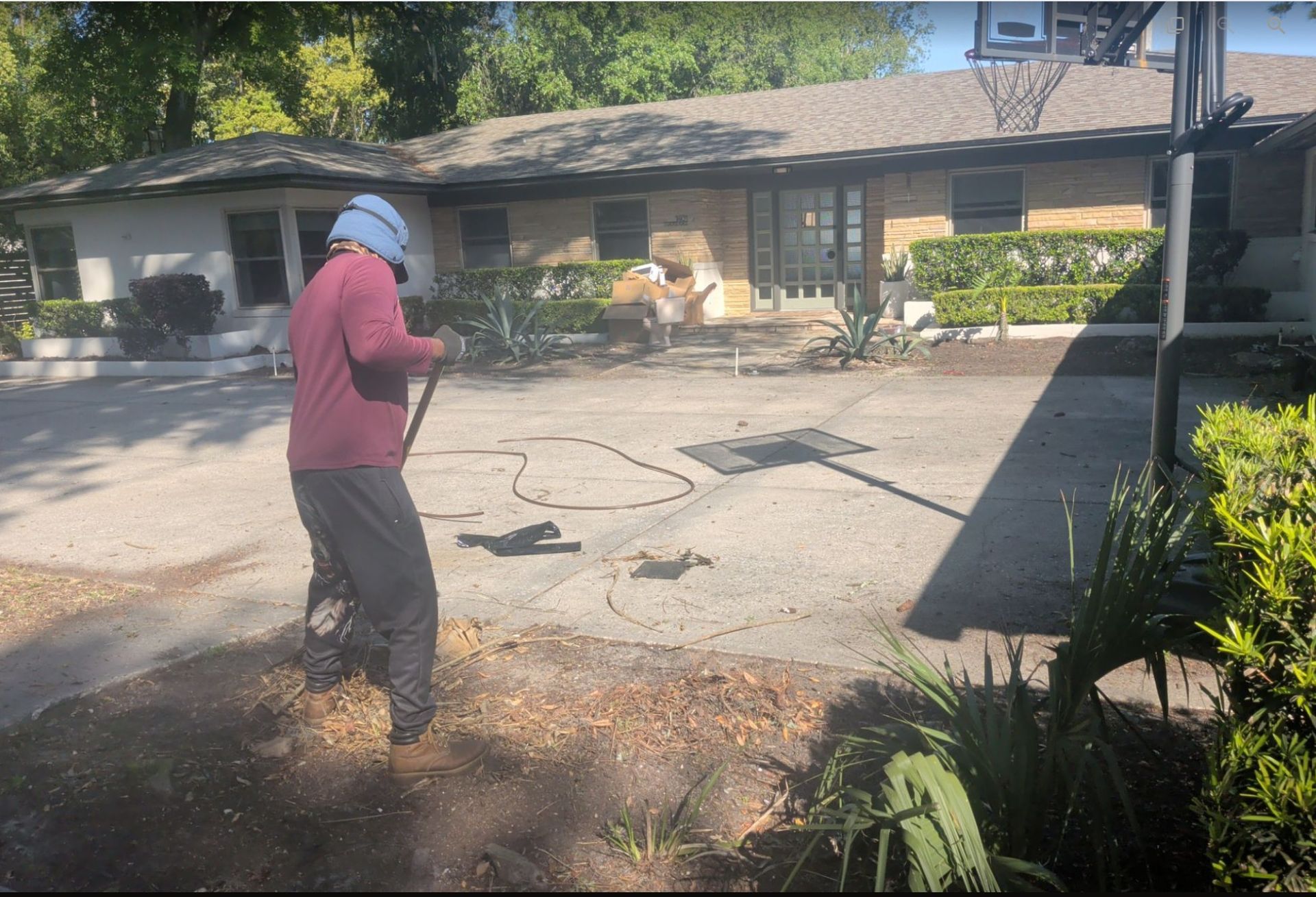 Person in a hat using a hose or pressure washer on a driveway in front of a house