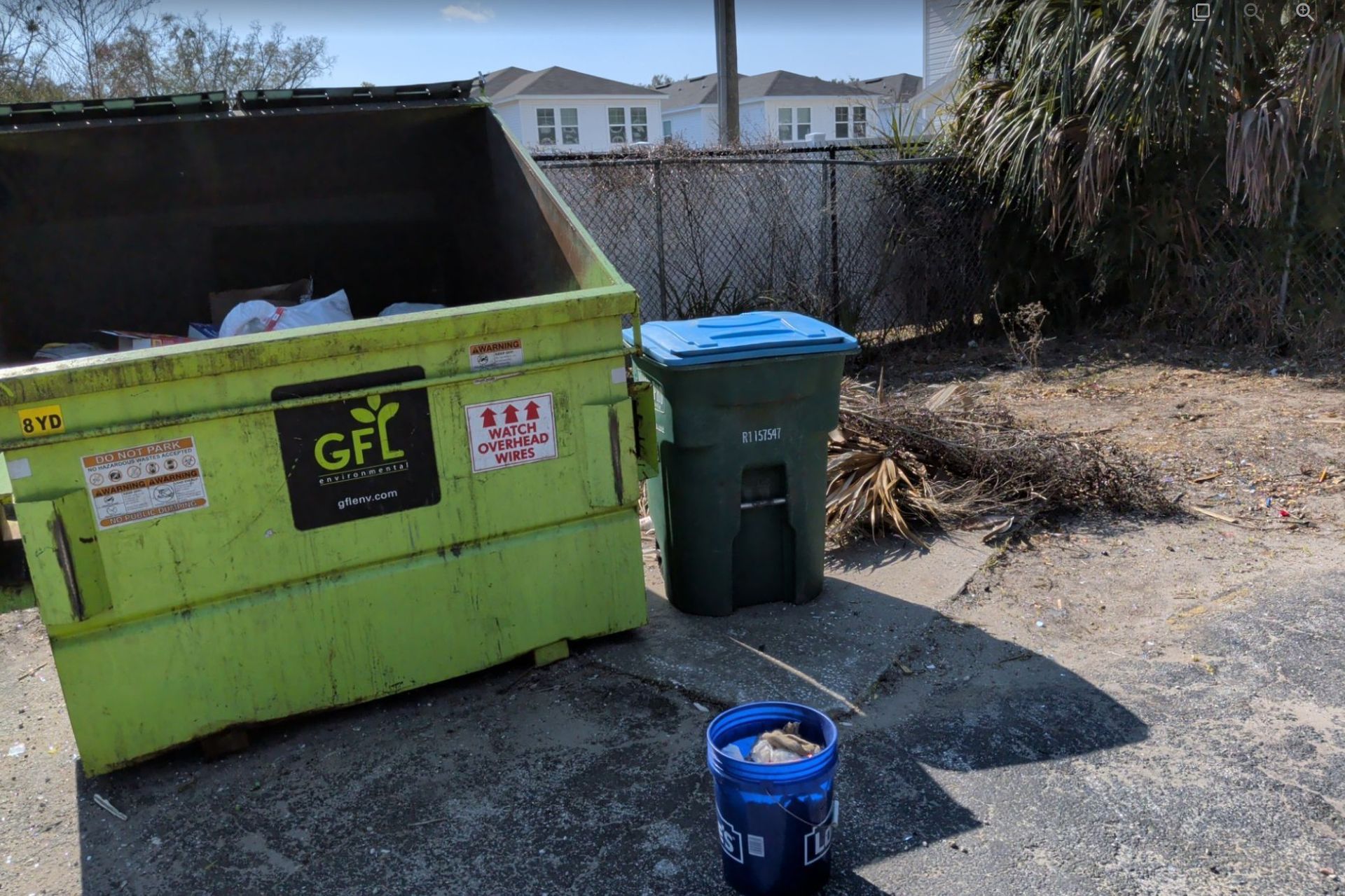 Green dumpster and trash bins beside a driveway with a blue bucket in front, sunny residential area