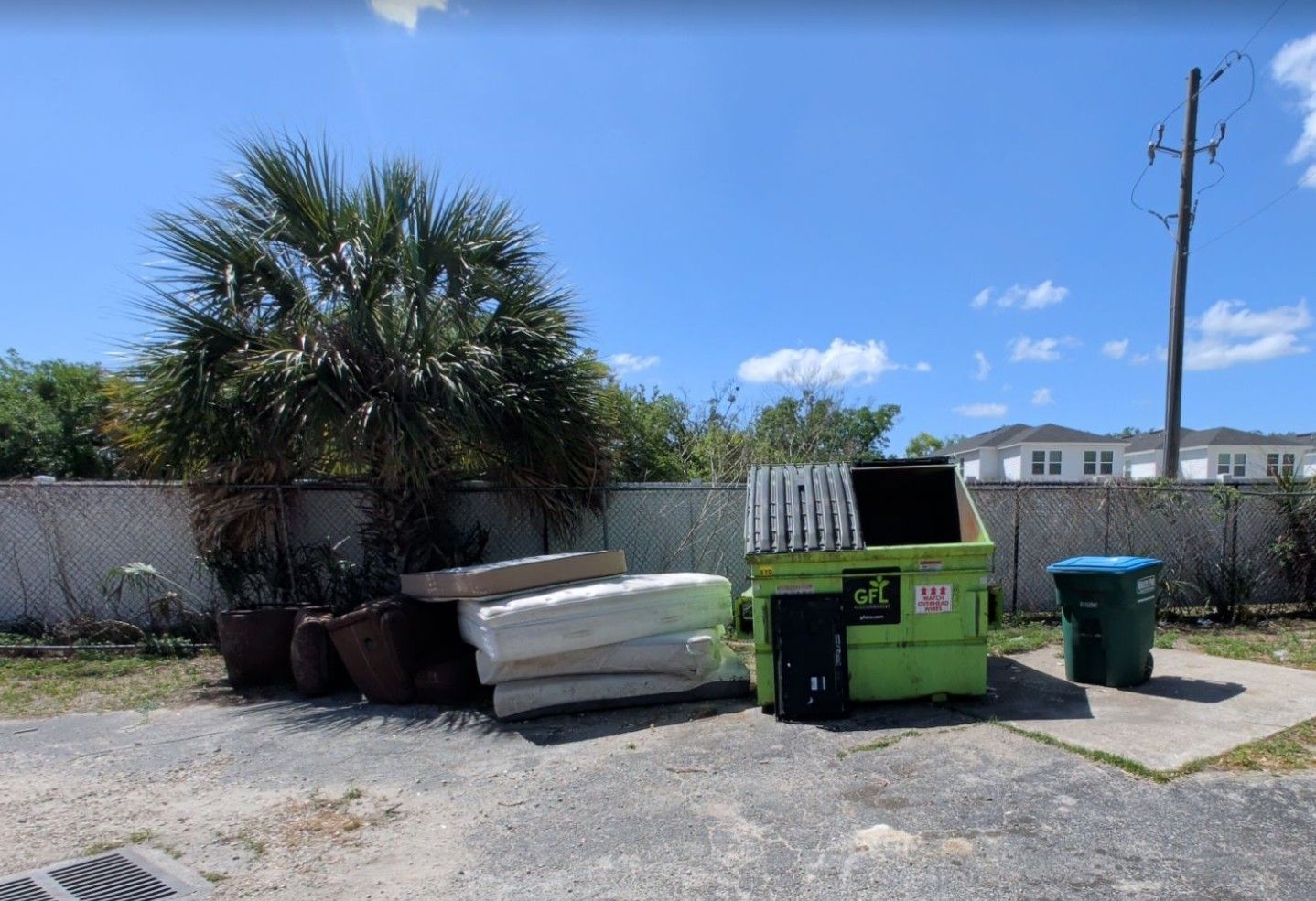 Dumpsters, a large white container, and trash bins beside a fence under a sunny blue sky.