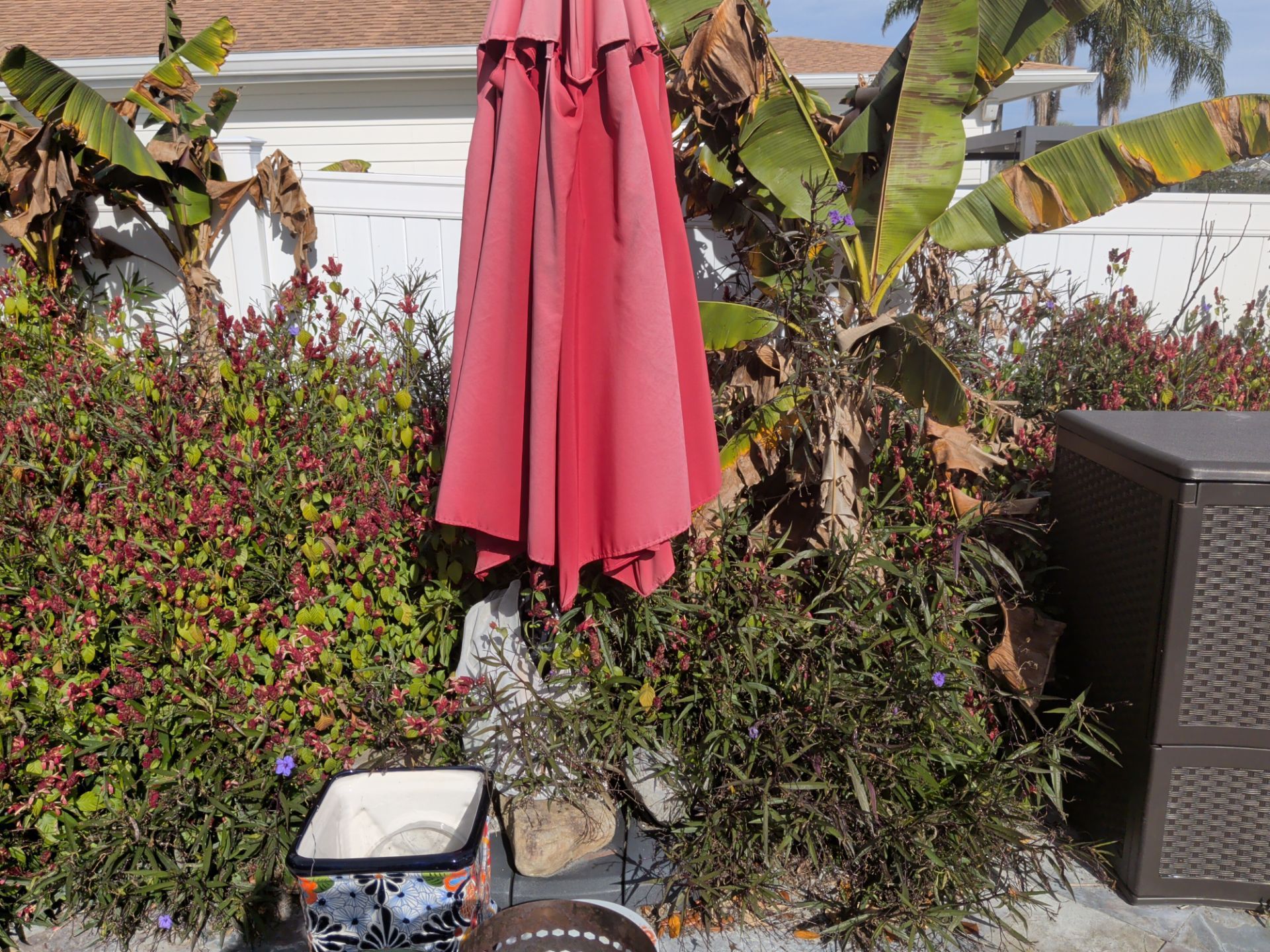 Backyard patio with a closed red umbrella, potted plants, and a grill beside a white wall.