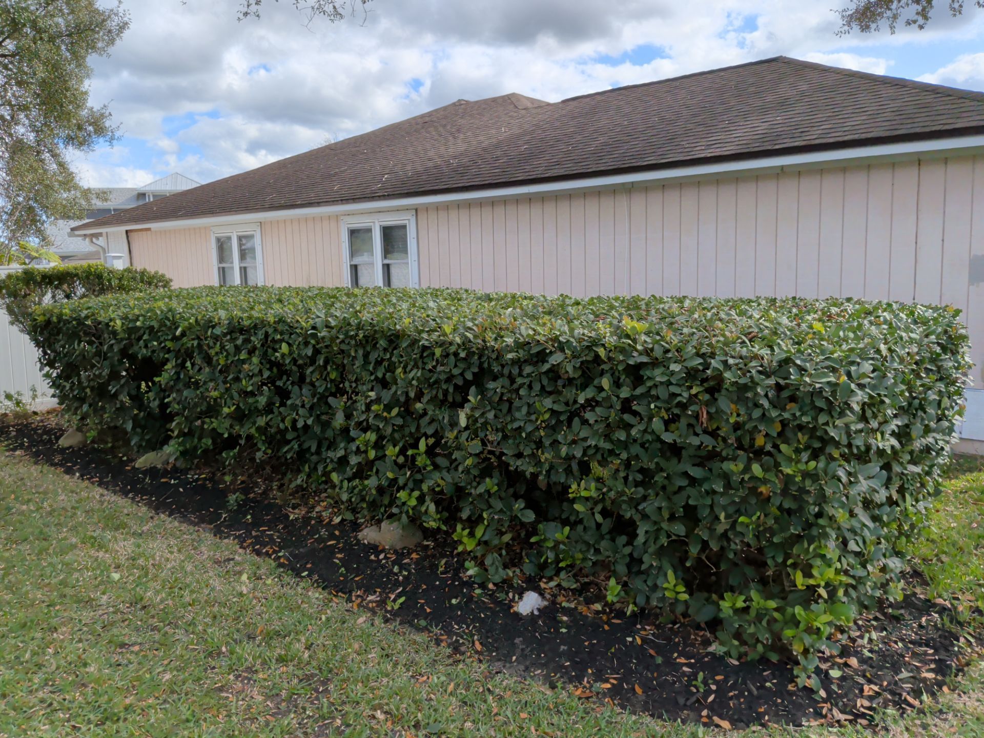 Green hedge in front of a light pink house with white windows and brown roof, set on a lawn.