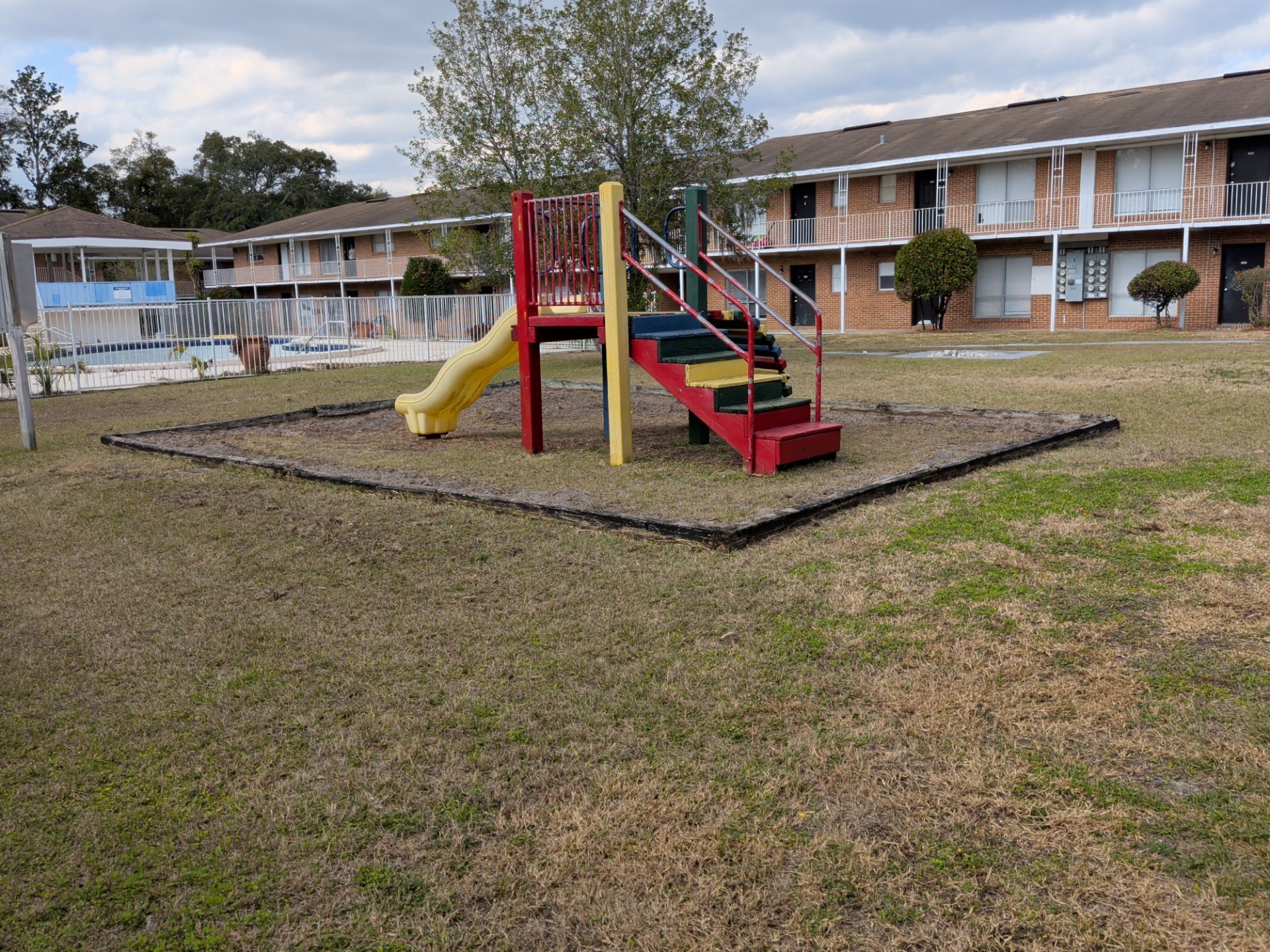 Playground with red and yellow slide, colorful stairs, grass, and apartment building in the background.