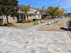 Paver sidewalk in front of two-story tan house, palm trees, blue sky.