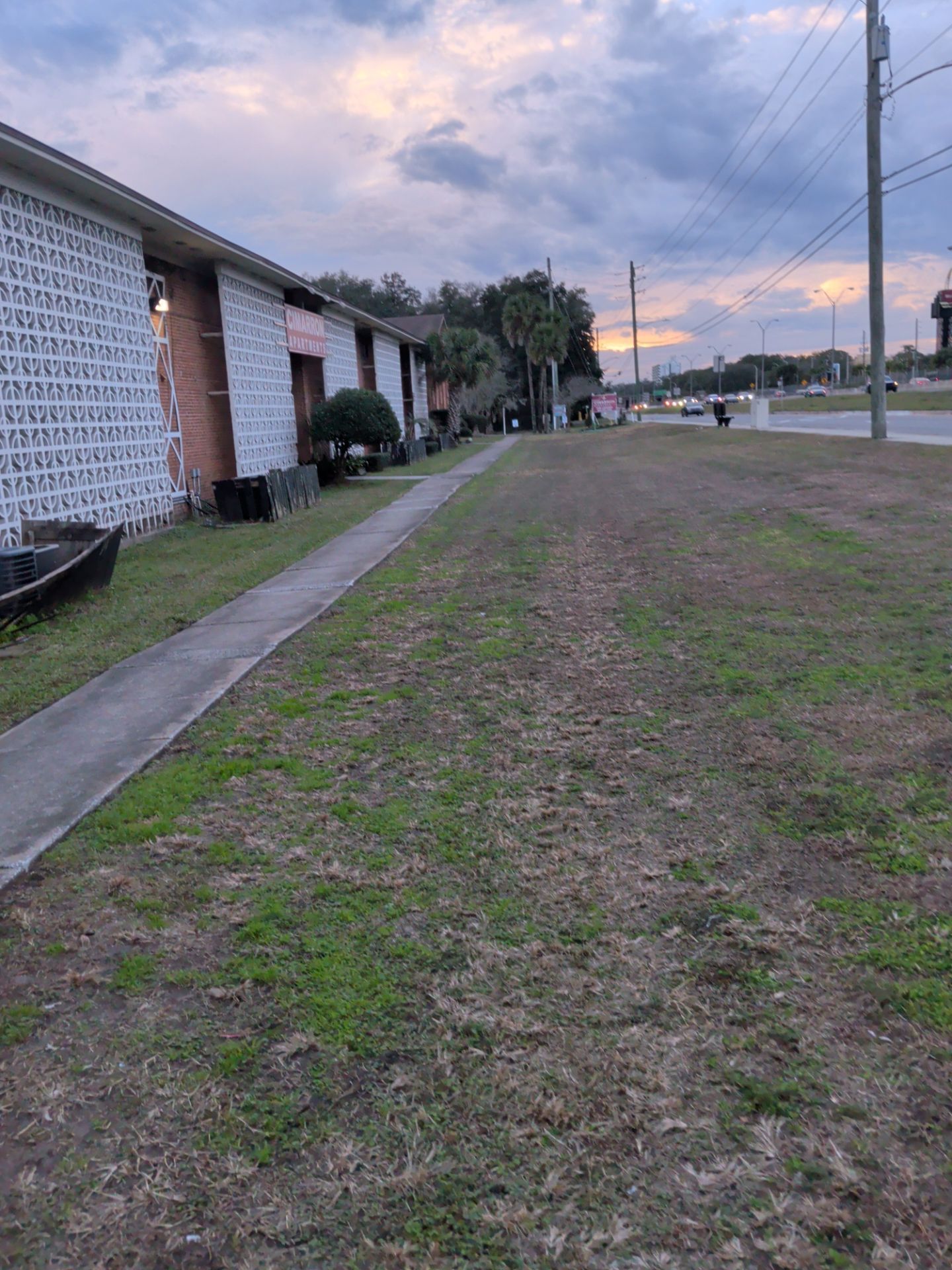 Apartment building exterior, sidewalk, and grassy area with a road under a cloudy sky.