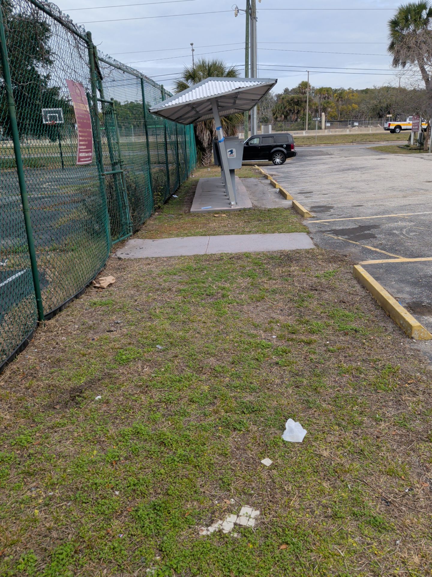 A bus shelter next to a chain-link fence and a parking lot. Cloudy sky.