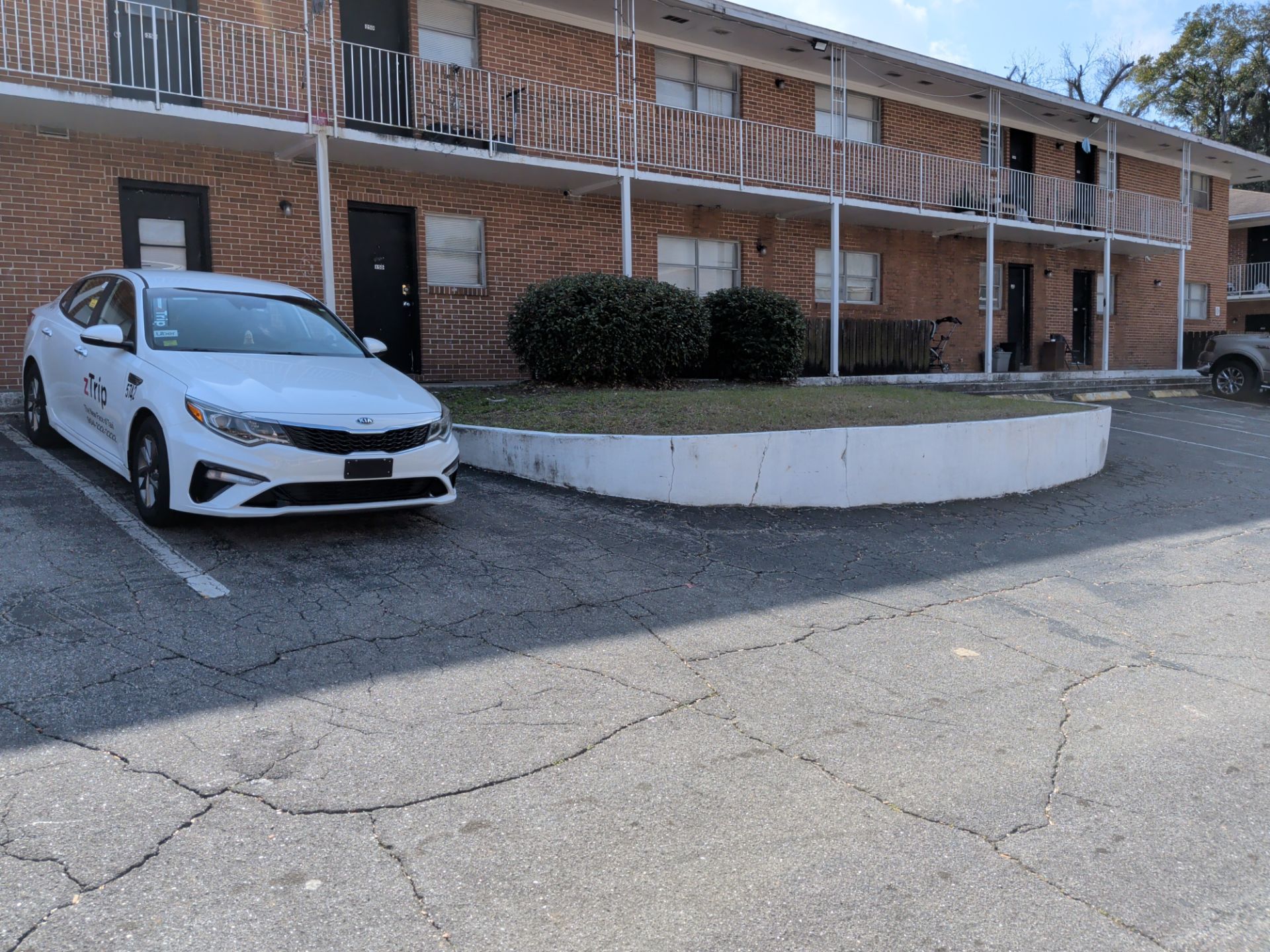 White car parked in front of a two-story brick apartment building. Sunny day, asphalt parking lot.
