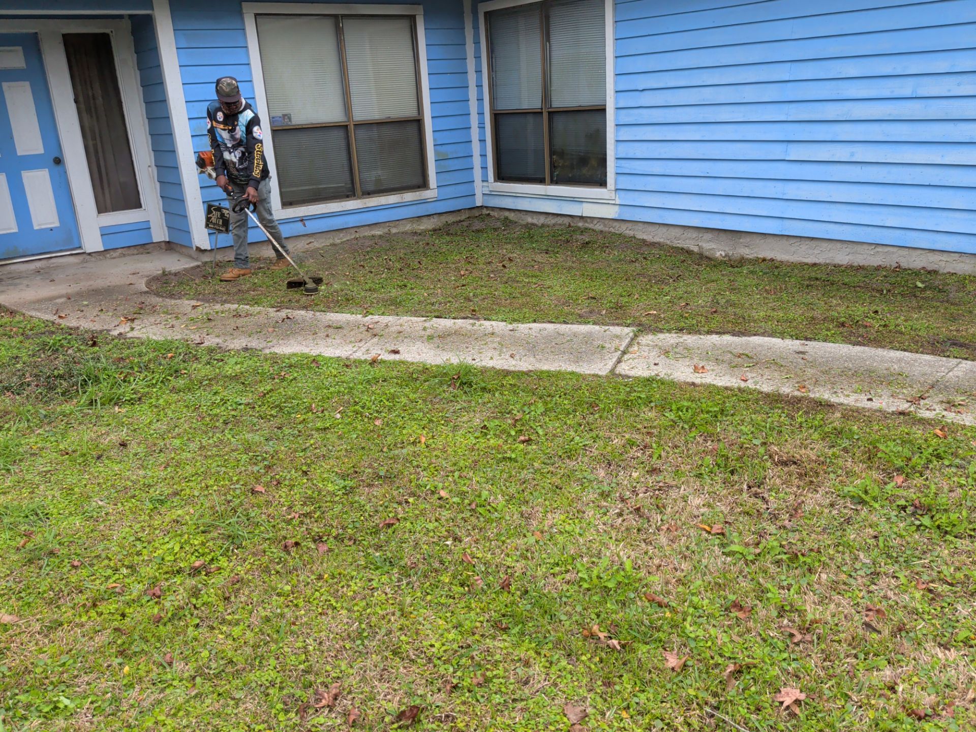 Person using a string trimmer on a lawn in front of a blue house.