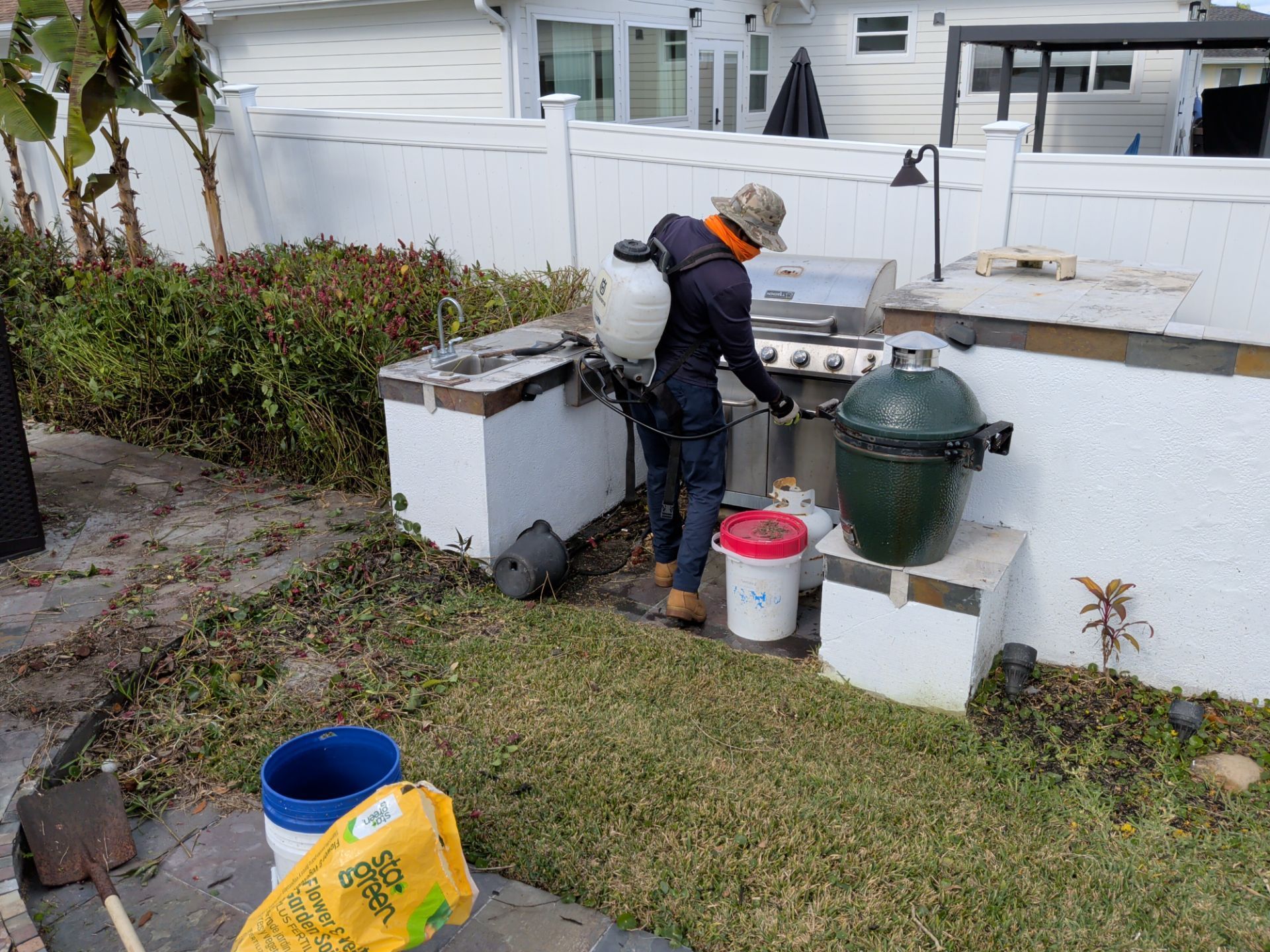 A person with a backpack sprayer near a built-in grill and Big Green Egg in a backyard.