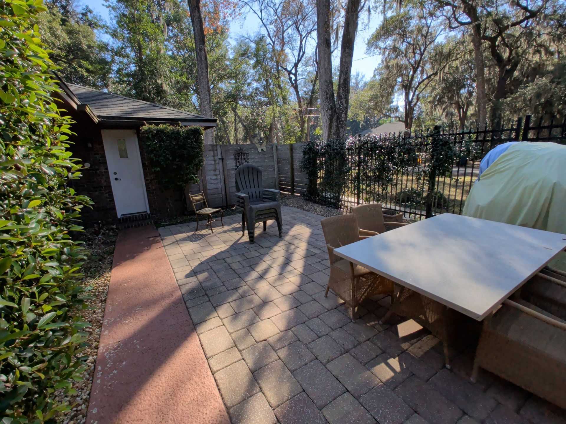Patio with brick pavers, table, chairs, fence, and a small building with a door. Sunny outdoor setting.