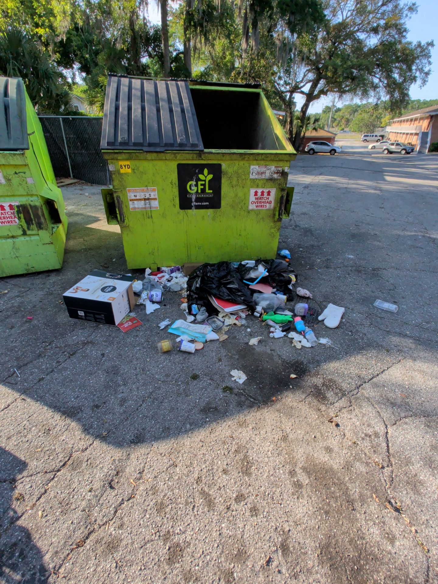 Green dumpster overflowing with scattered trash on pavement in a parking lot