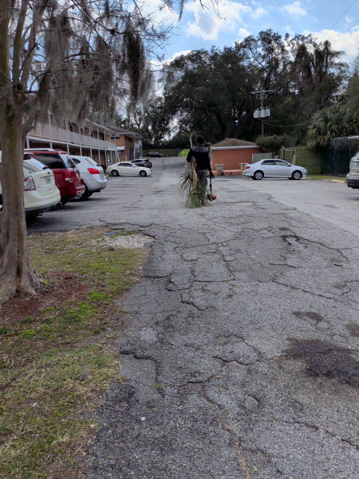 Person pushing a bicycle on a cracked asphalt parking lot, next to parked cars and a low building.