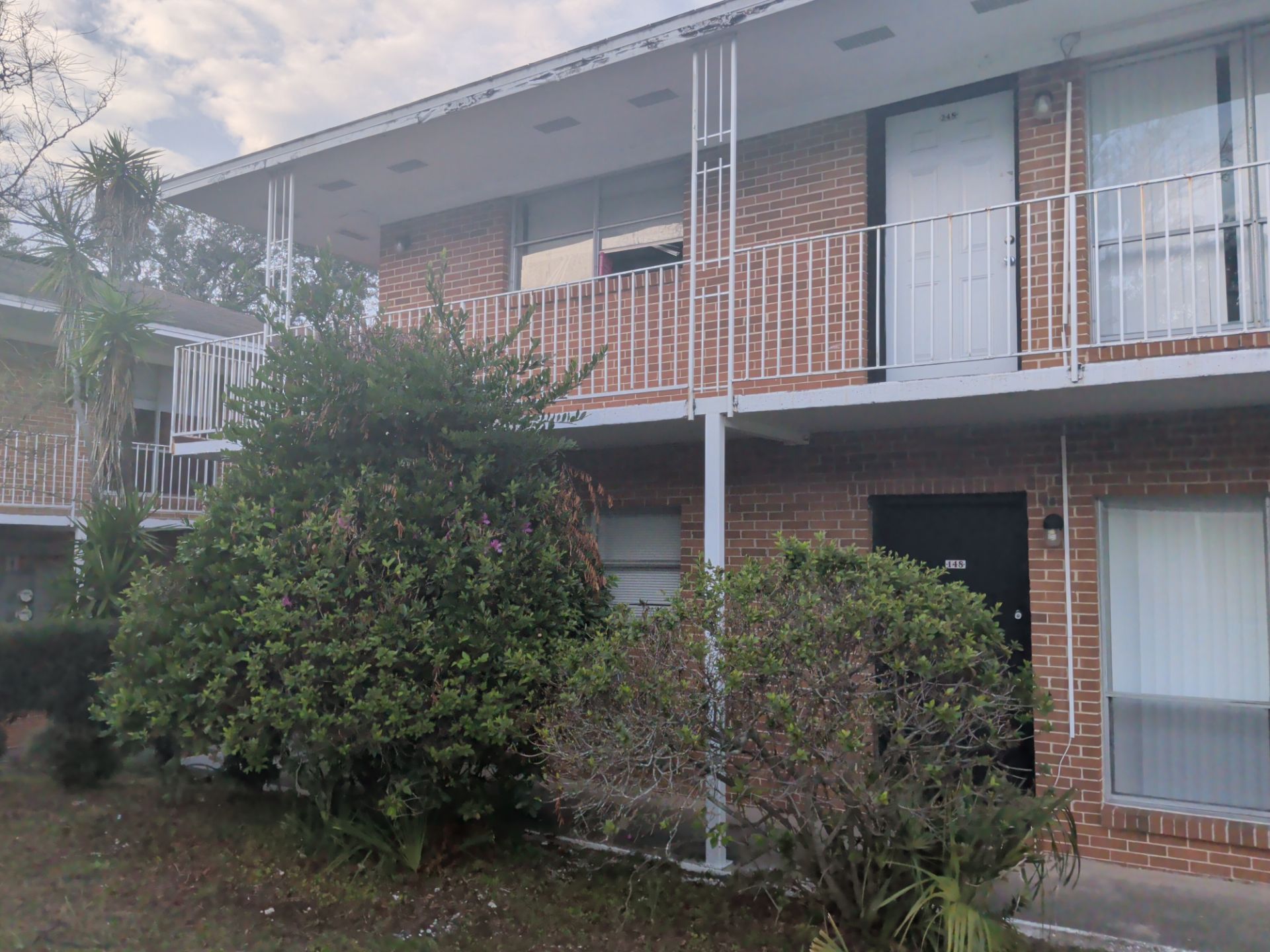 Two-story brick apartment building with white balconies and overgrown bushes. Cloudy sky.