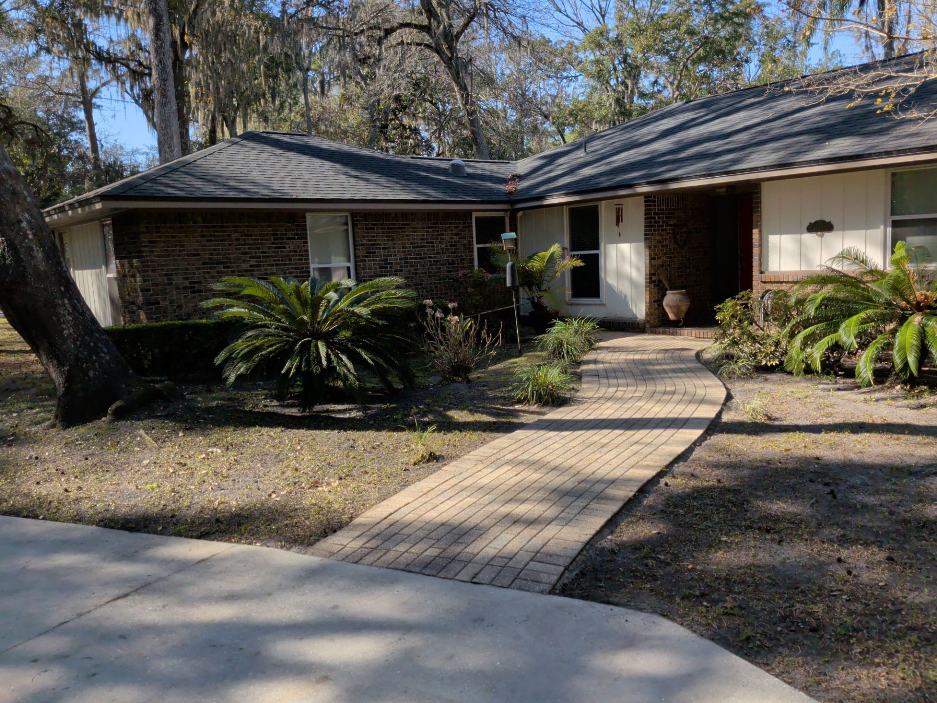 Brick walkway leads to a one-story house with dark shingles, brick, and light-colored walls; sunny day.