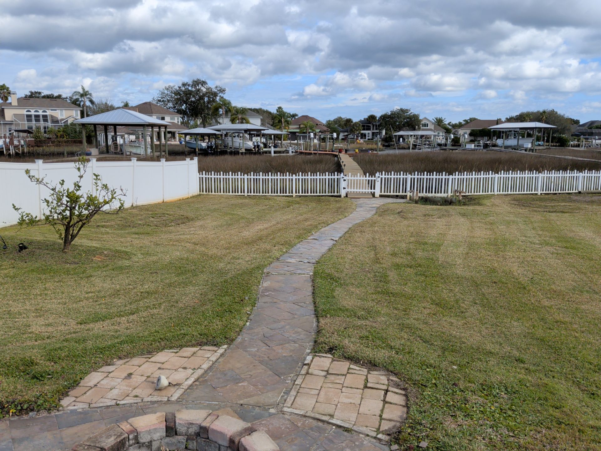 Grassy backyard with stone path leading to white picket fence and waterfront houses under a cloudy sky.