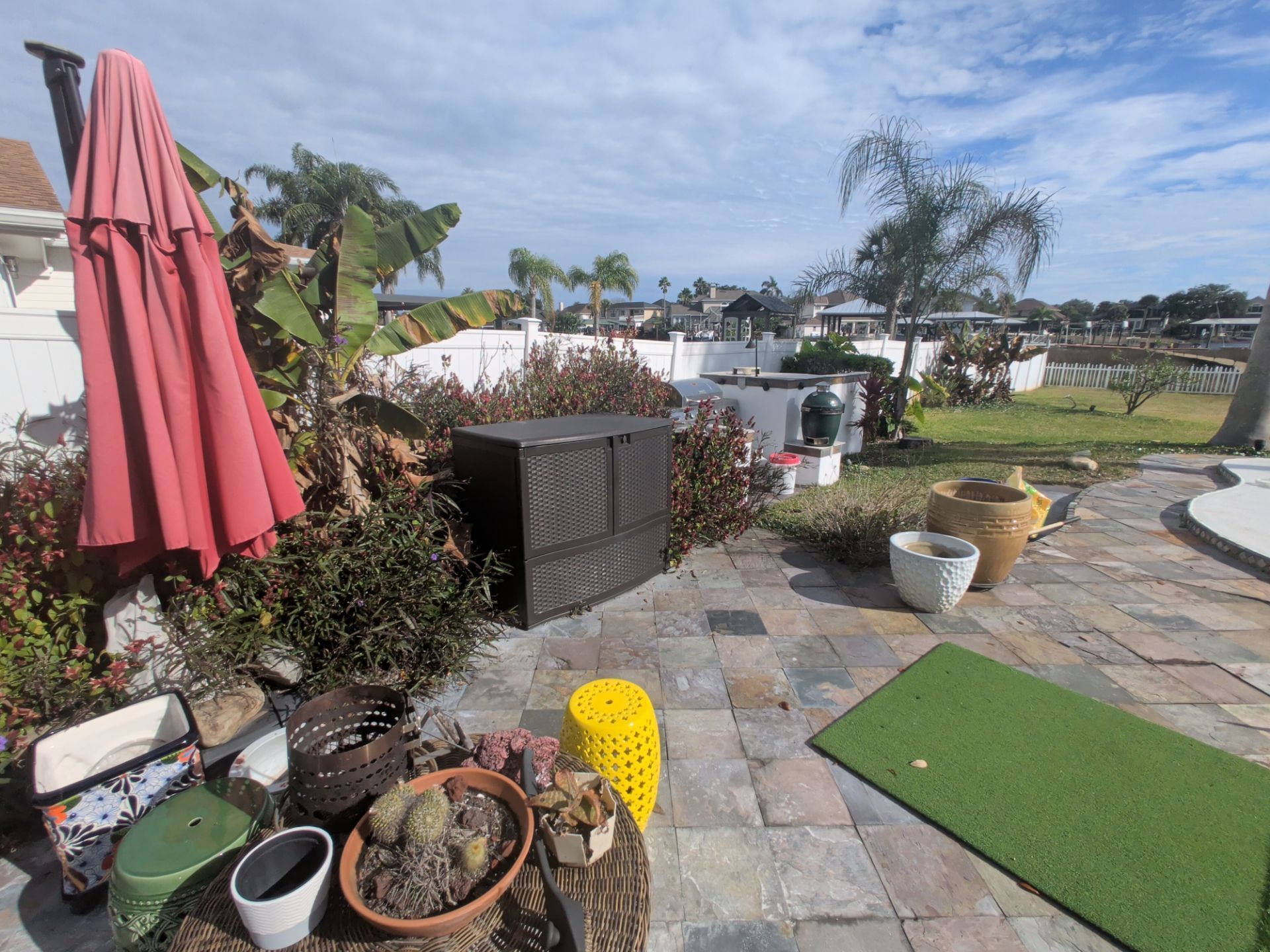 Backyard with brick patio, plants, umbrella, and putting green under cloudy sky.