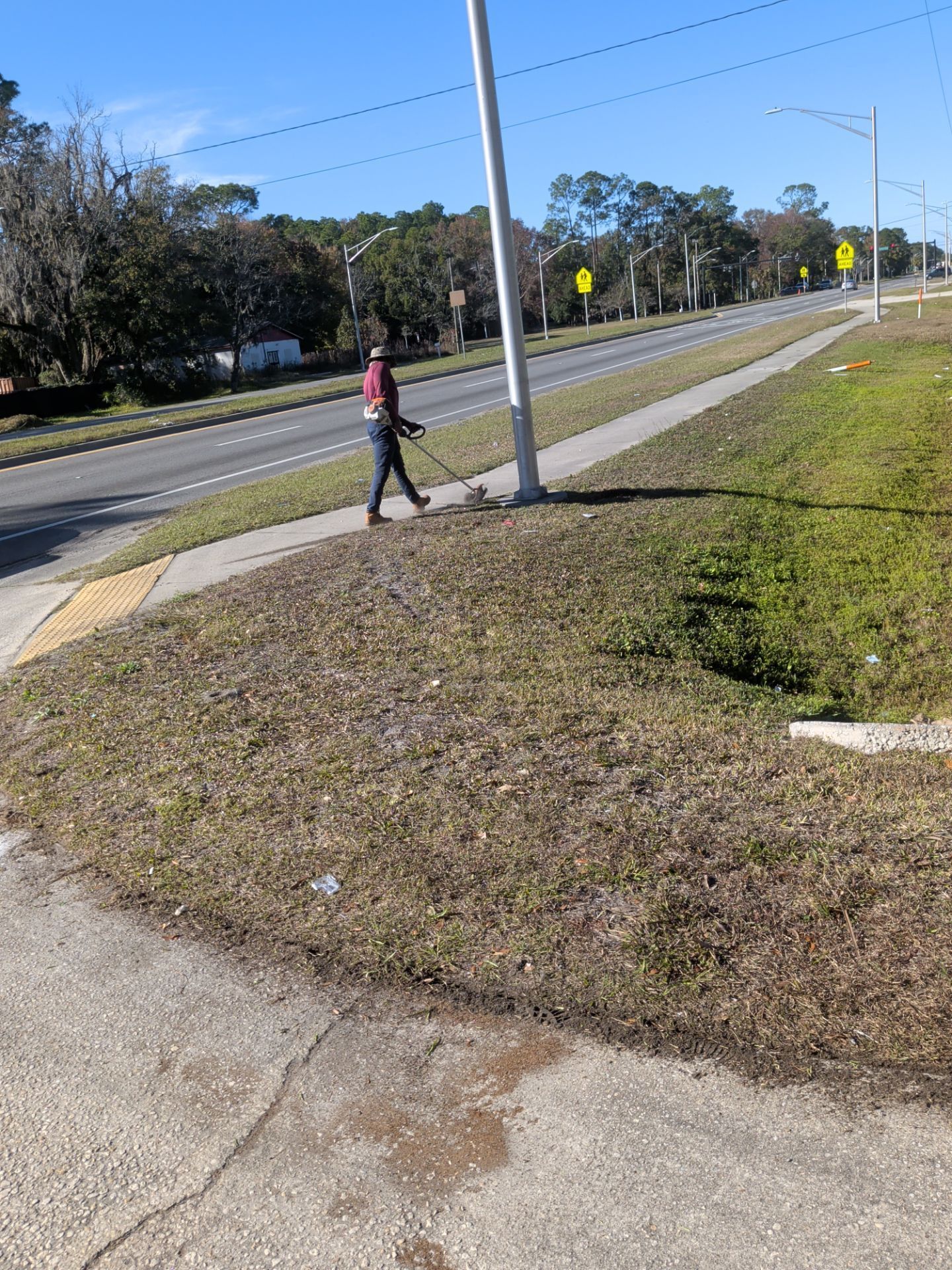 Person walking on sidewalk along road, using a tool. Clear, sunny day.