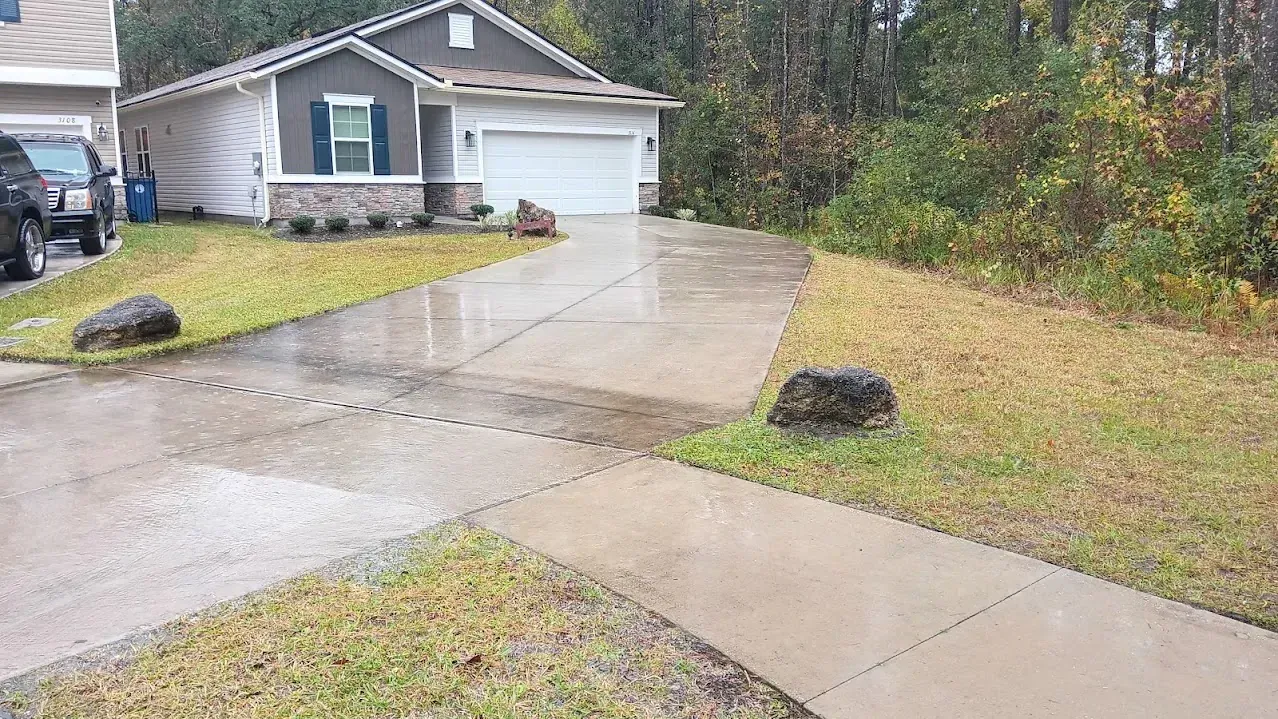Wet driveway leading to a beige house with a closed garage door; two large rocks in grassy areas.