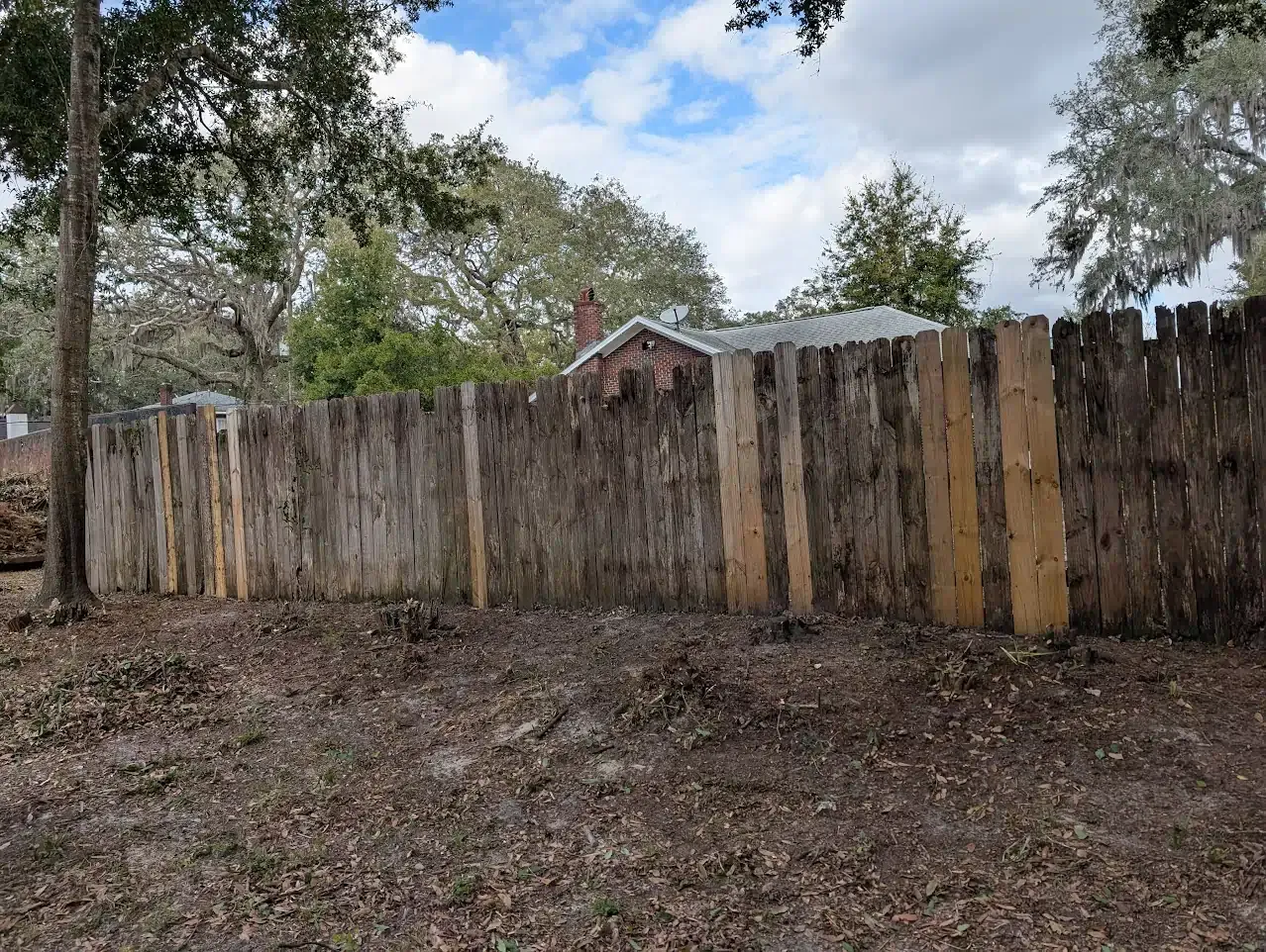 Weathered wooden fence in a yard, trees in the background, a house with a brick chimney visible.
