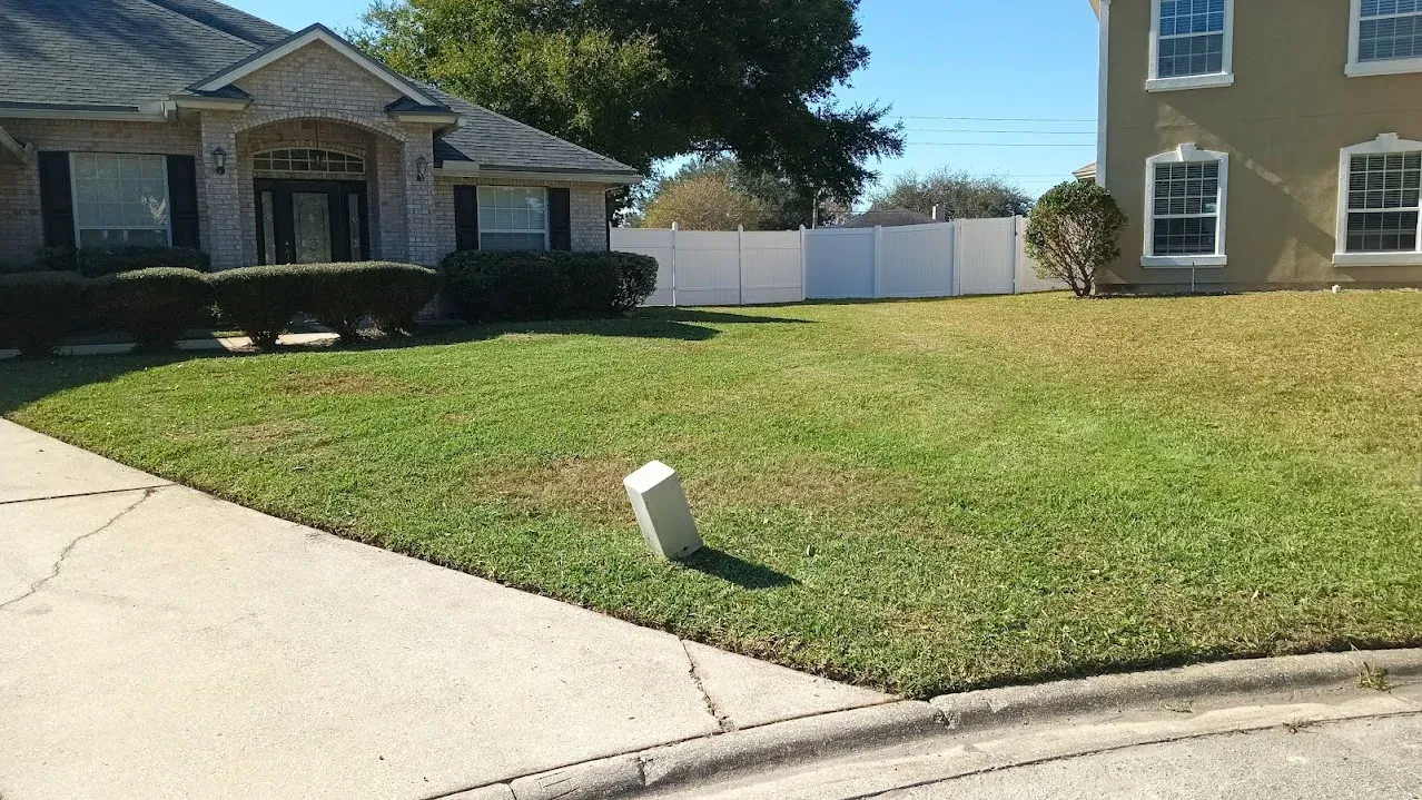 Suburban house with patchy green lawn, a white fence, and a sunny sky.