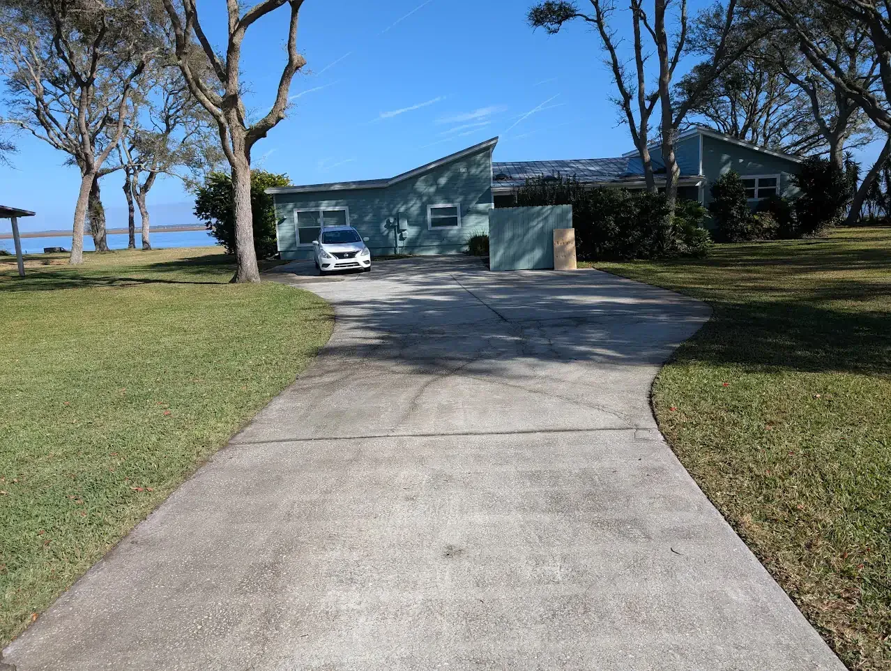 A teal house with a slanted roof sits near a body of water. A concrete driveway leads up to it.