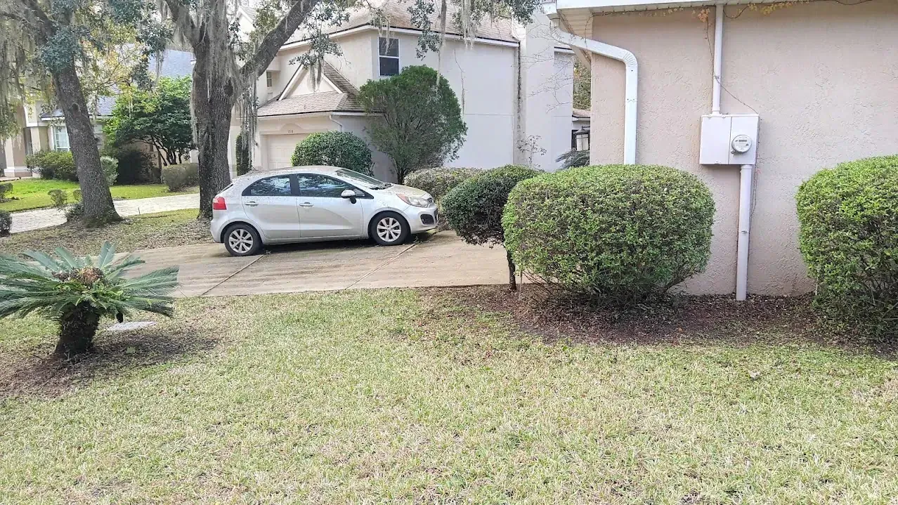 Silver car parked on a driveway with dry leaves; house and shrubs in the yard.