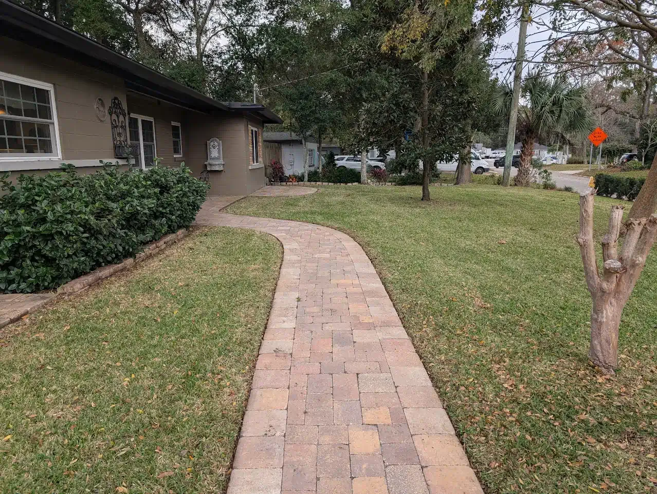 Brick pathway curves toward a house, flanked by green grass and a small tree.