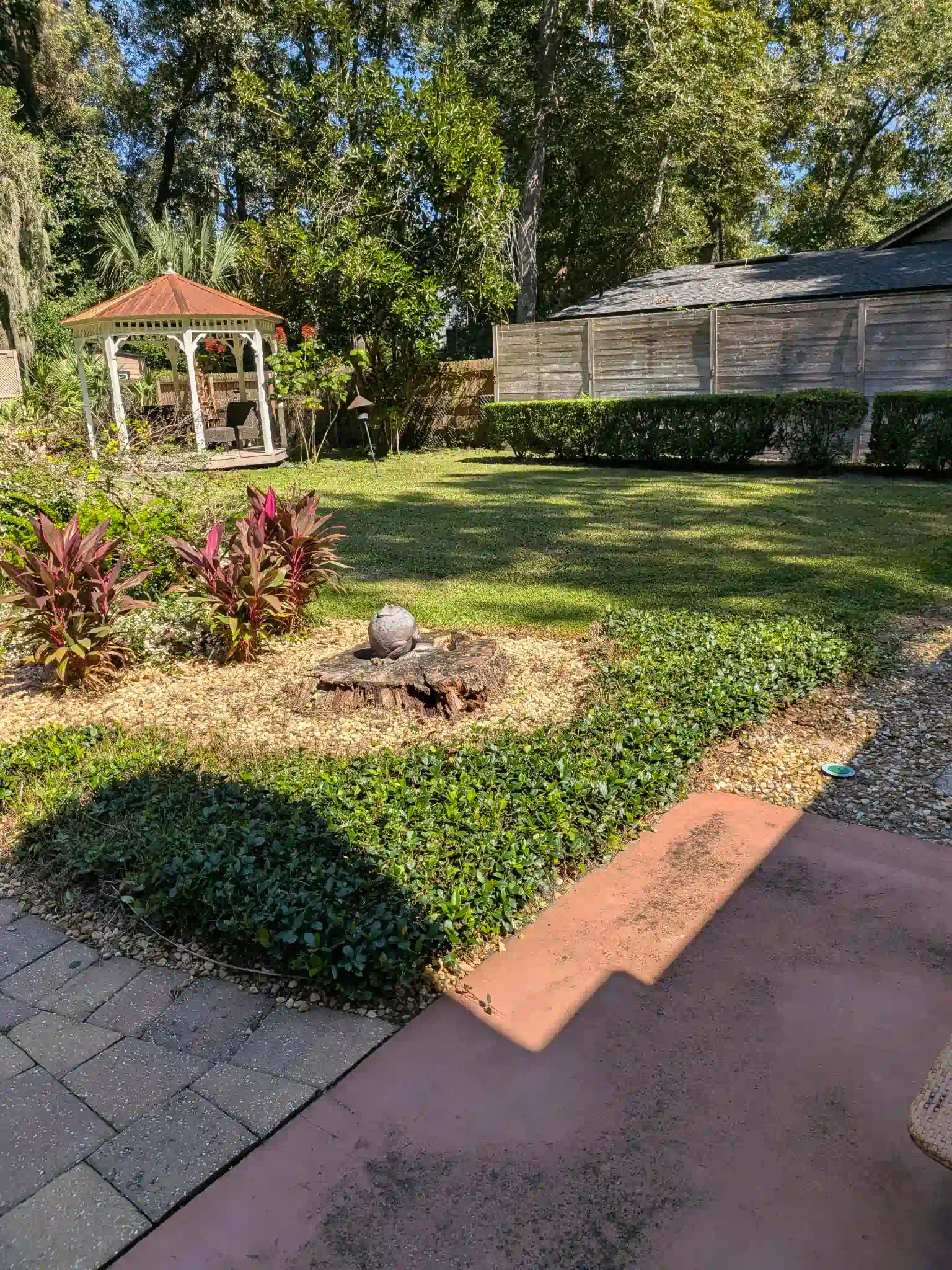 Backyard with a gazebo, lawn, and garden beds. Red and green plants, wooden fence, and brick patio.