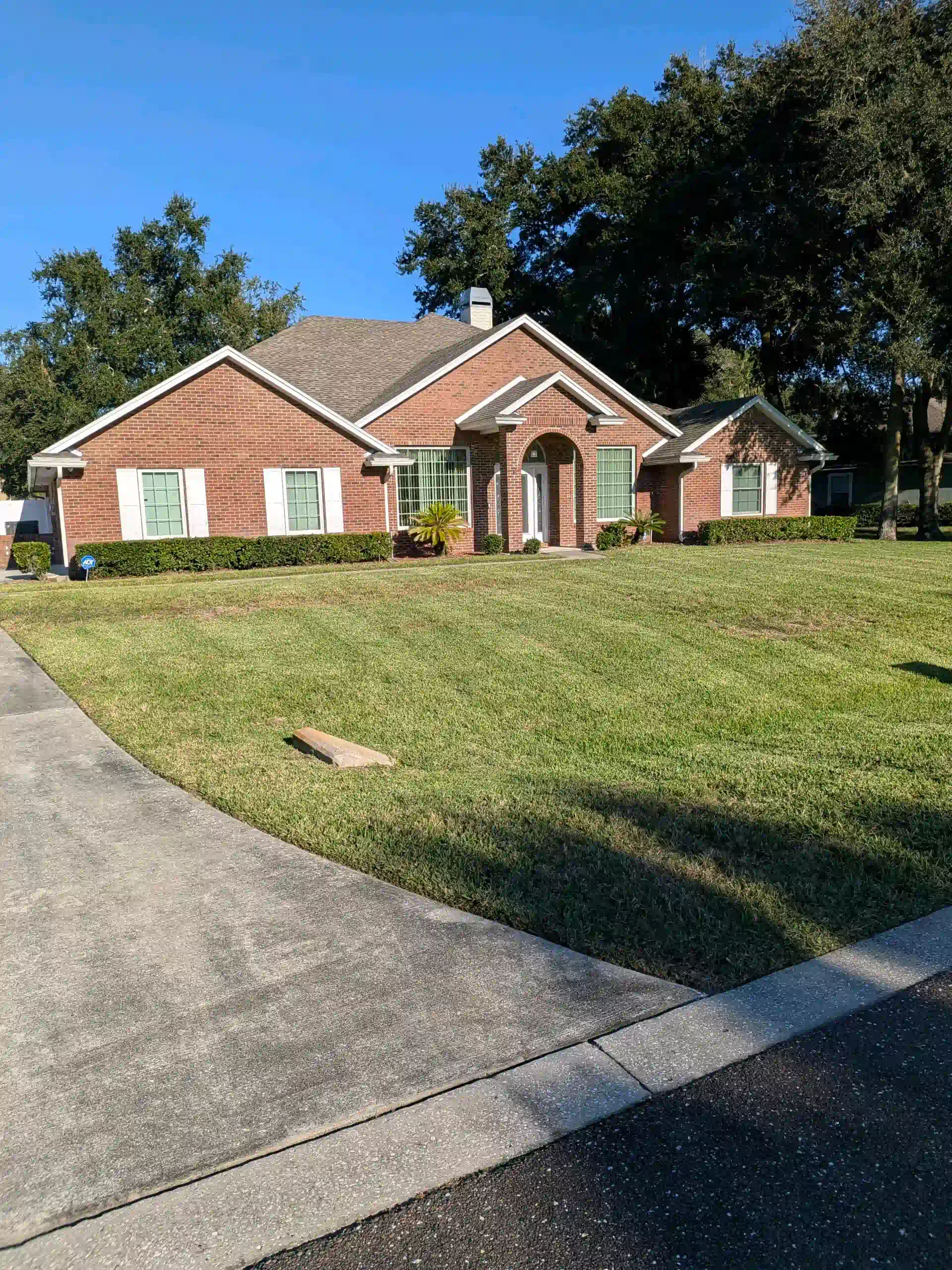 Brick house with a grassy lawn, sidewalk, and trees under a blue sky.