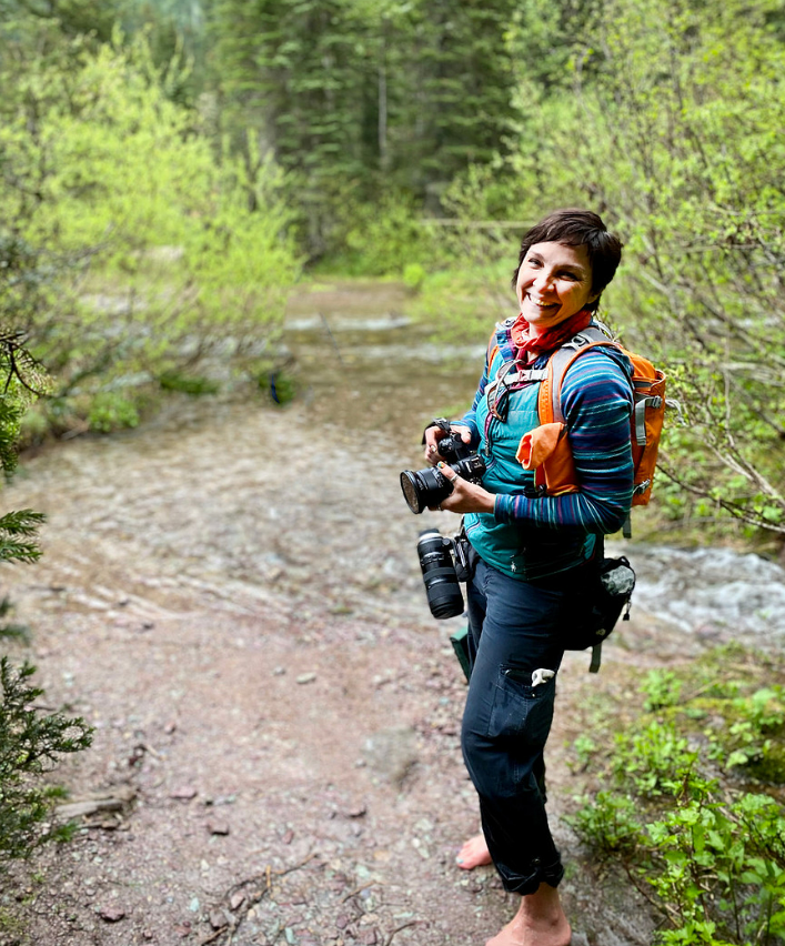Woman with cameras, barefoot, standing on path by stream in forest. She wears vest and backpack.