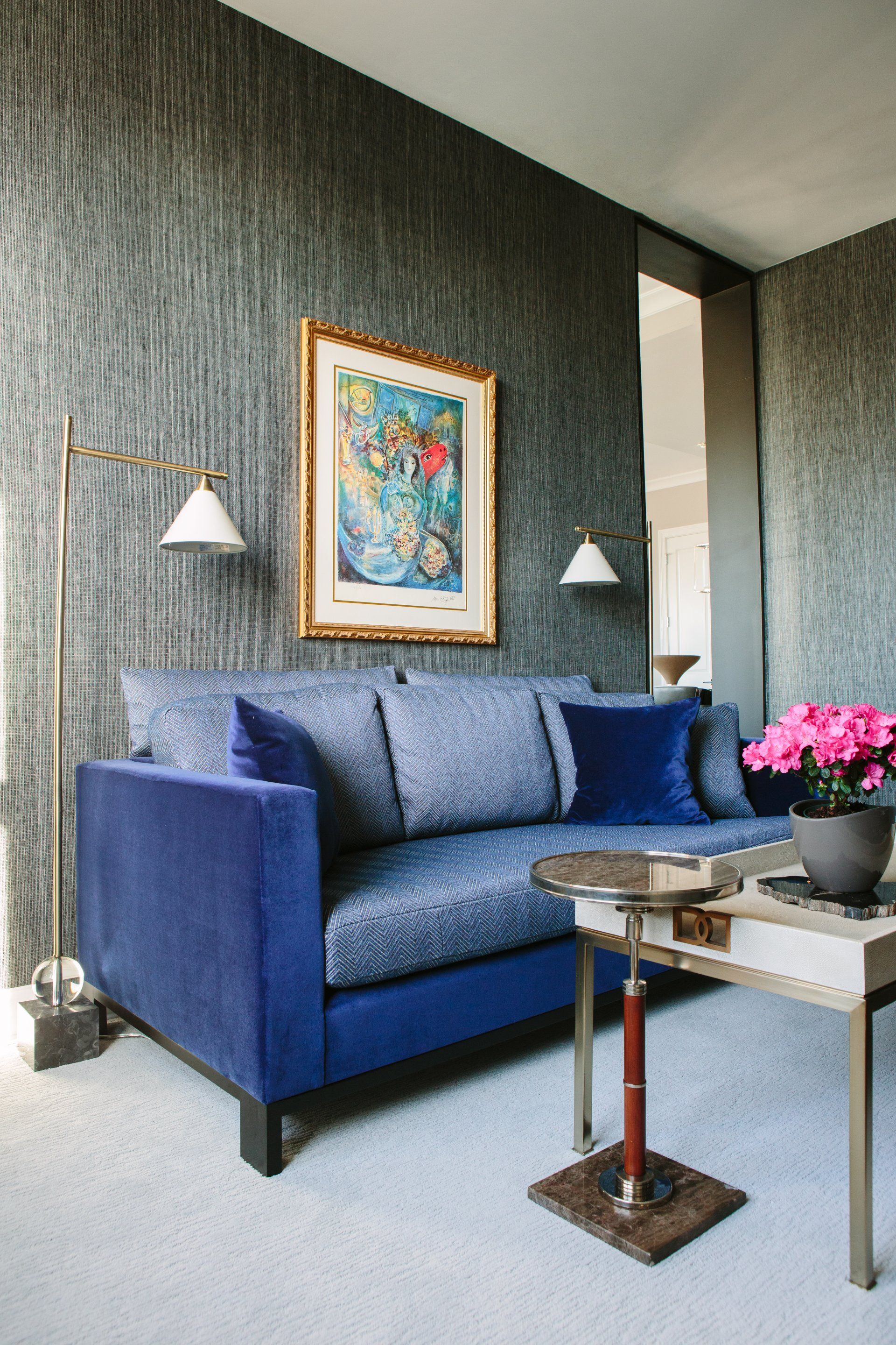 A living room with a blue couch , table , lamp and painting on the wall.