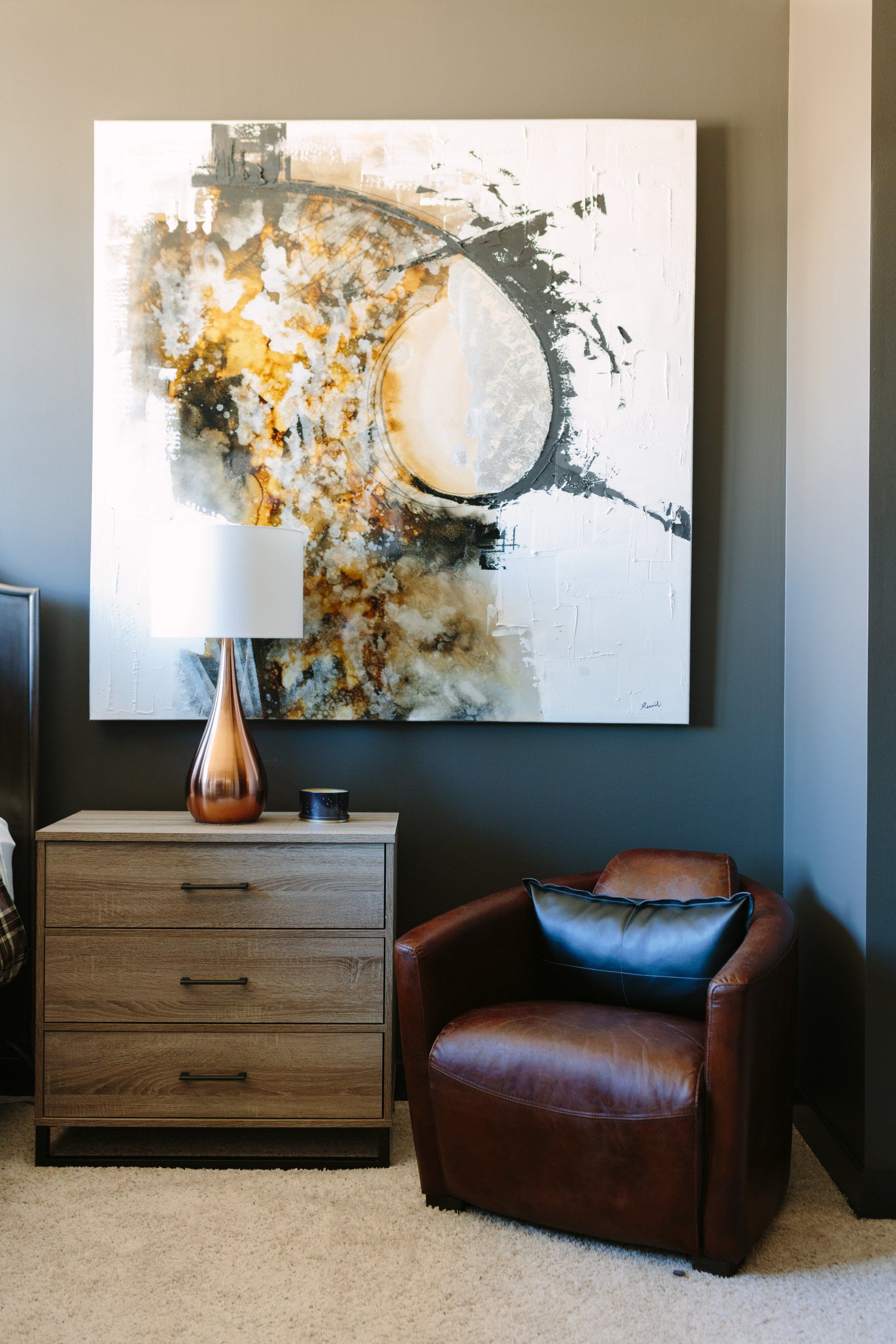 A bedroom with a chair , dresser , lamp and painting on the wall.