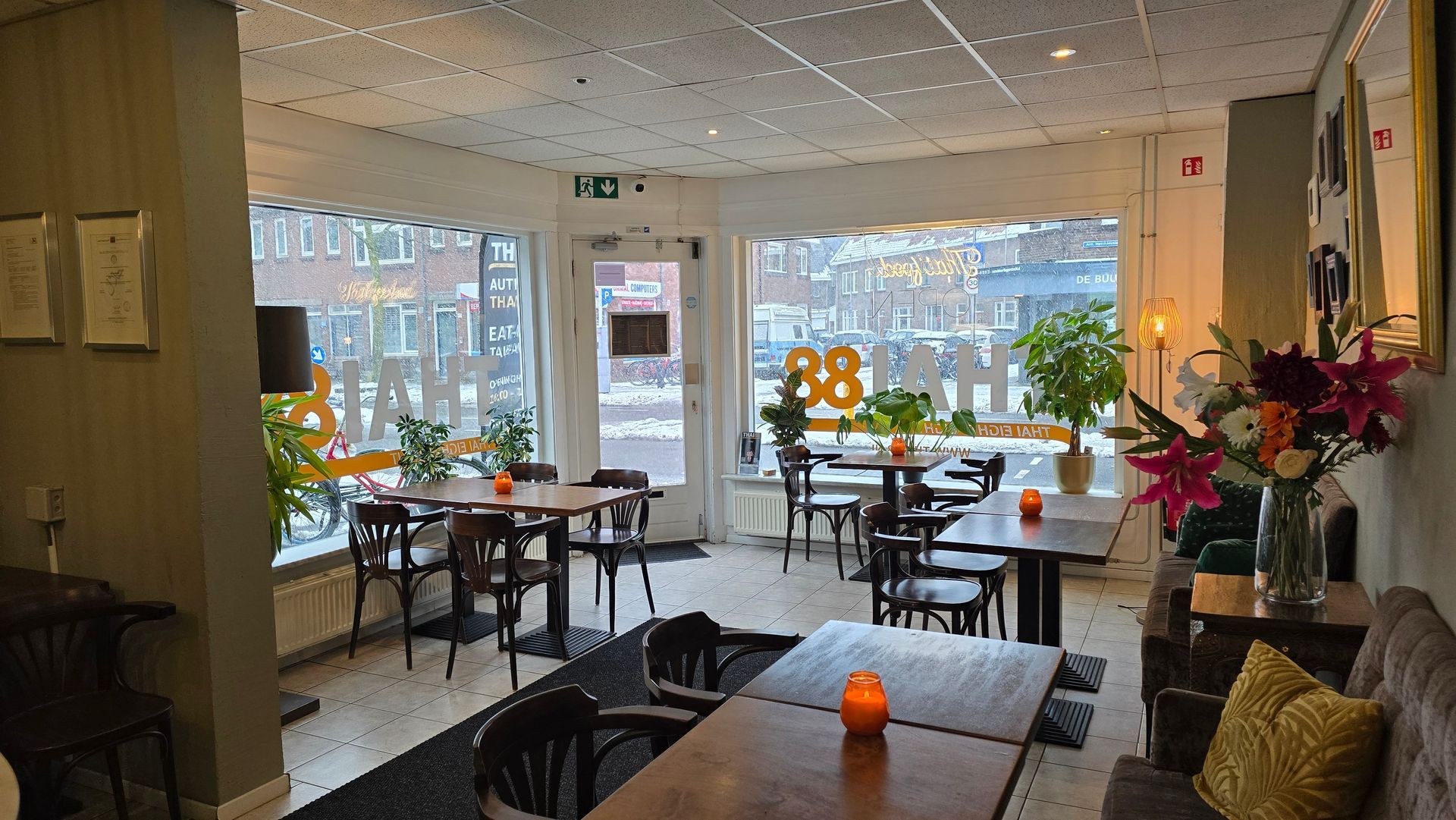 Interieurfoto van een gezellig, warm verlicht restaurant met meerdere eettafels, houten stoelen en grote ramen aan de straatkant.